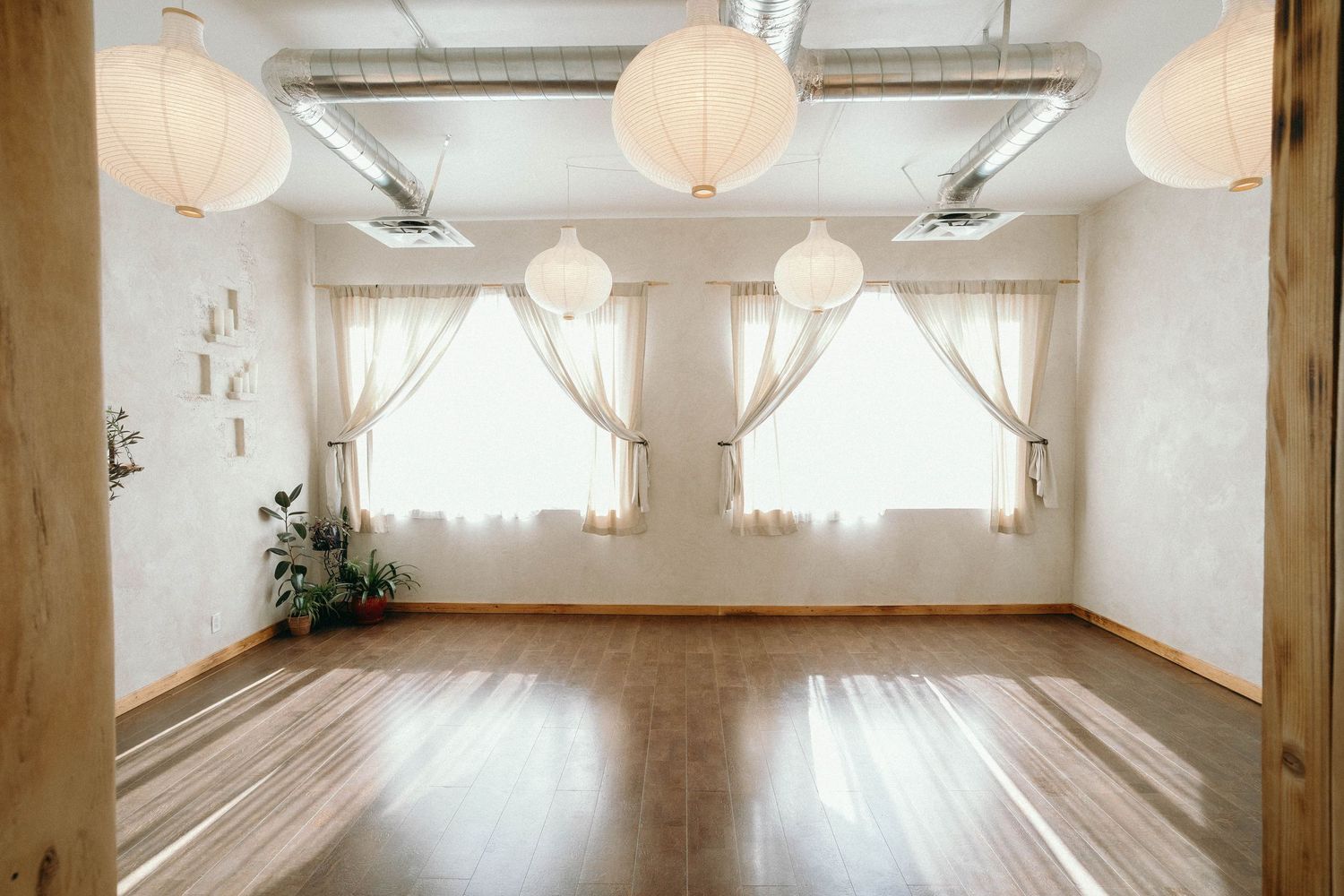 Bright yoga studio with hardwood floors, white paper lanterns, sheer curtains, and large windows creating a serene atmosphere.