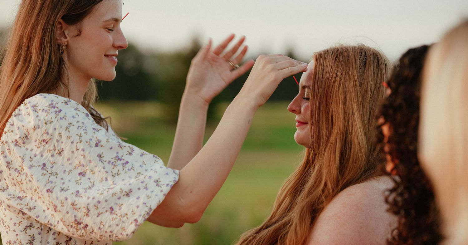 Two young women with long hair share a joyful moment outdoors, one gently touching the other's face.