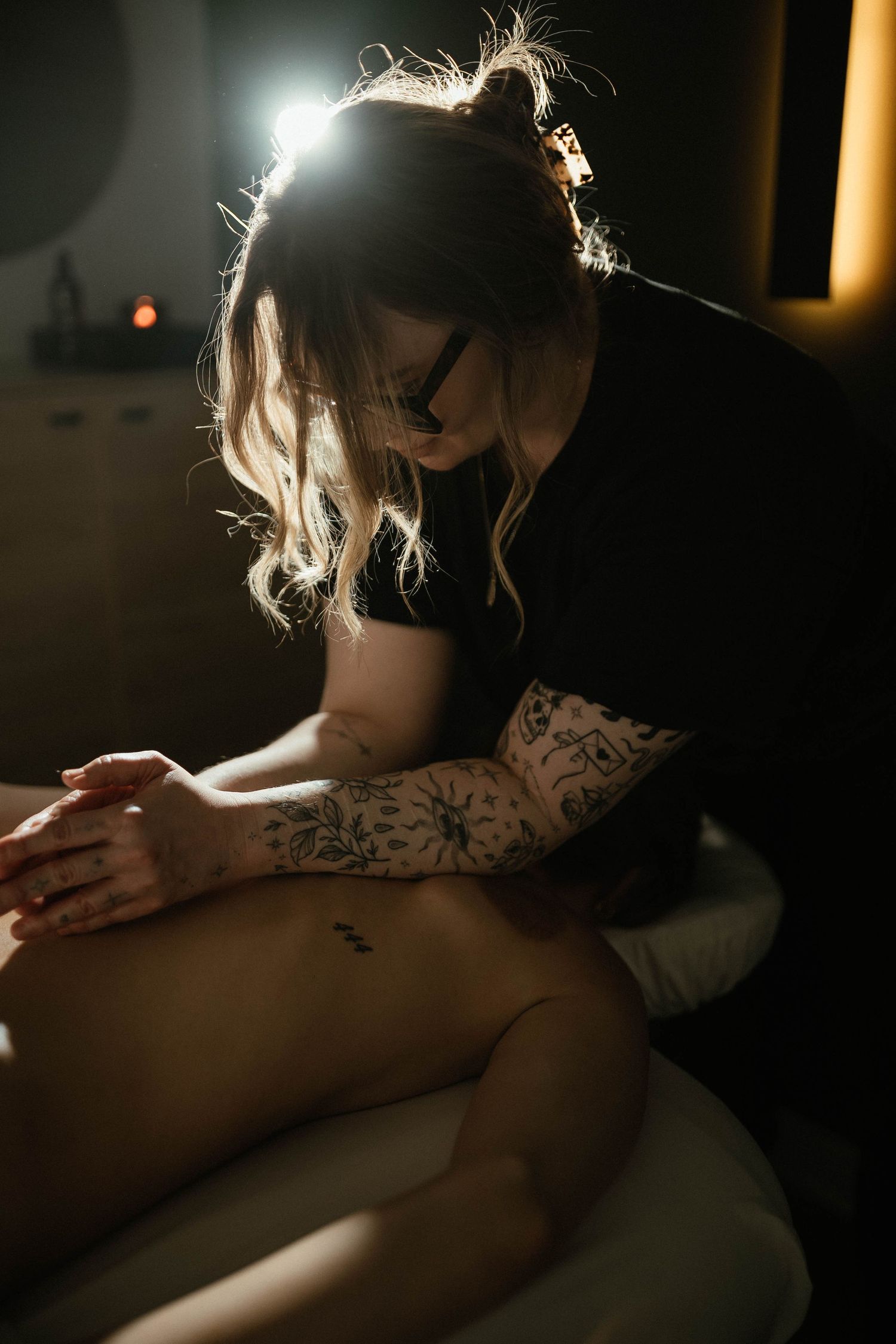 Tattoo artist with sleeve tattoos works on client in dimly lit studio, wearing black shirt and glasses.