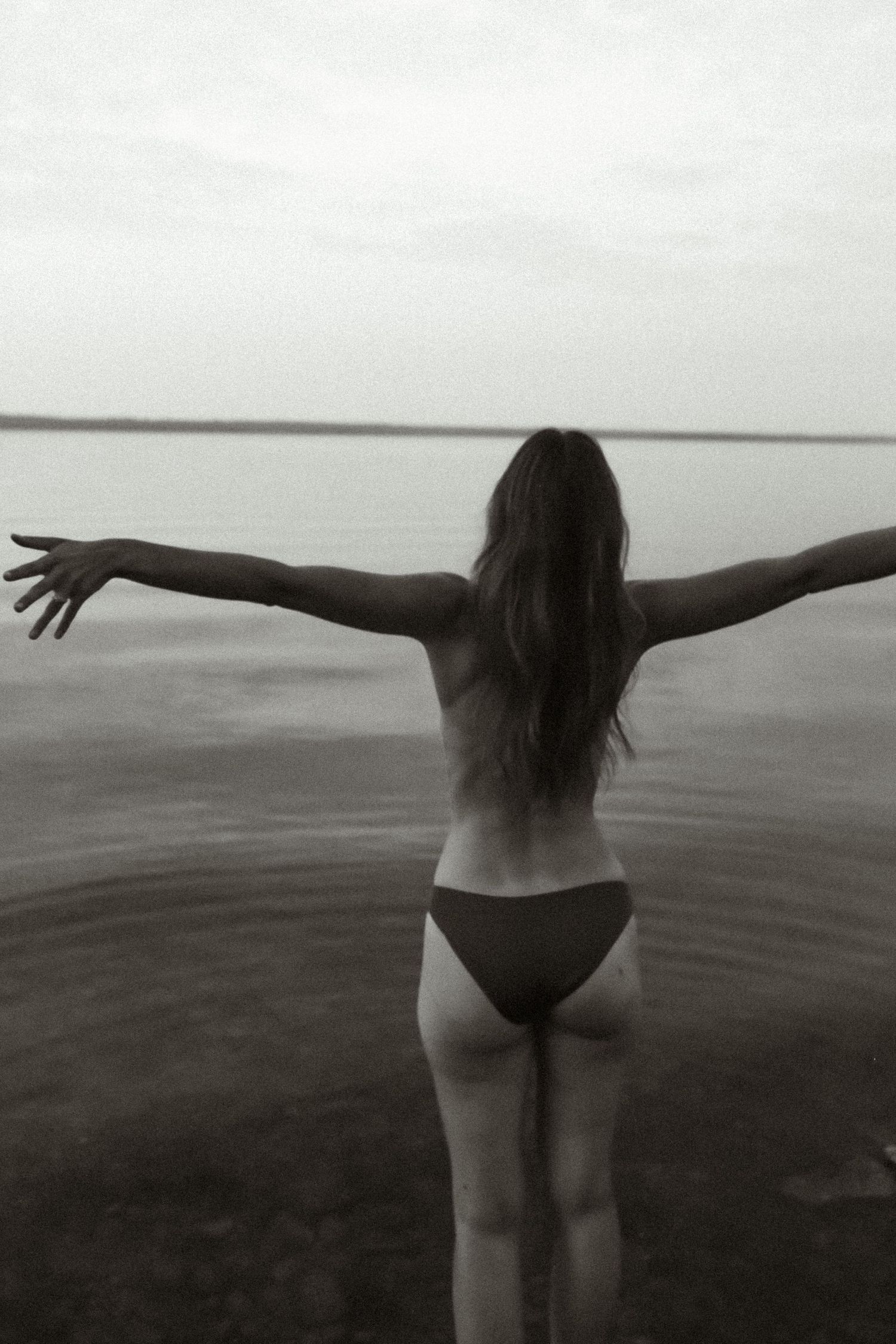 Black and white photo of woman with arms outstretched facing a lake, wearing bikini bottoms, shot from behind.