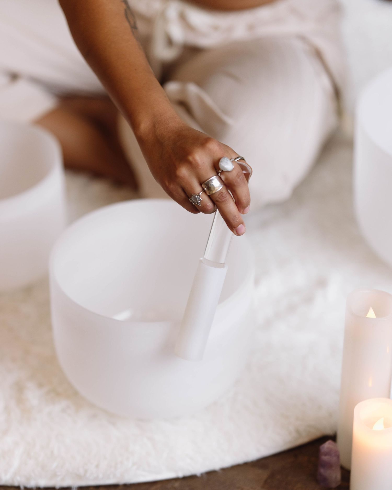 Person playing a white crystal singing bowl with a mallet, surrounded by candles in a serene meditation setting.