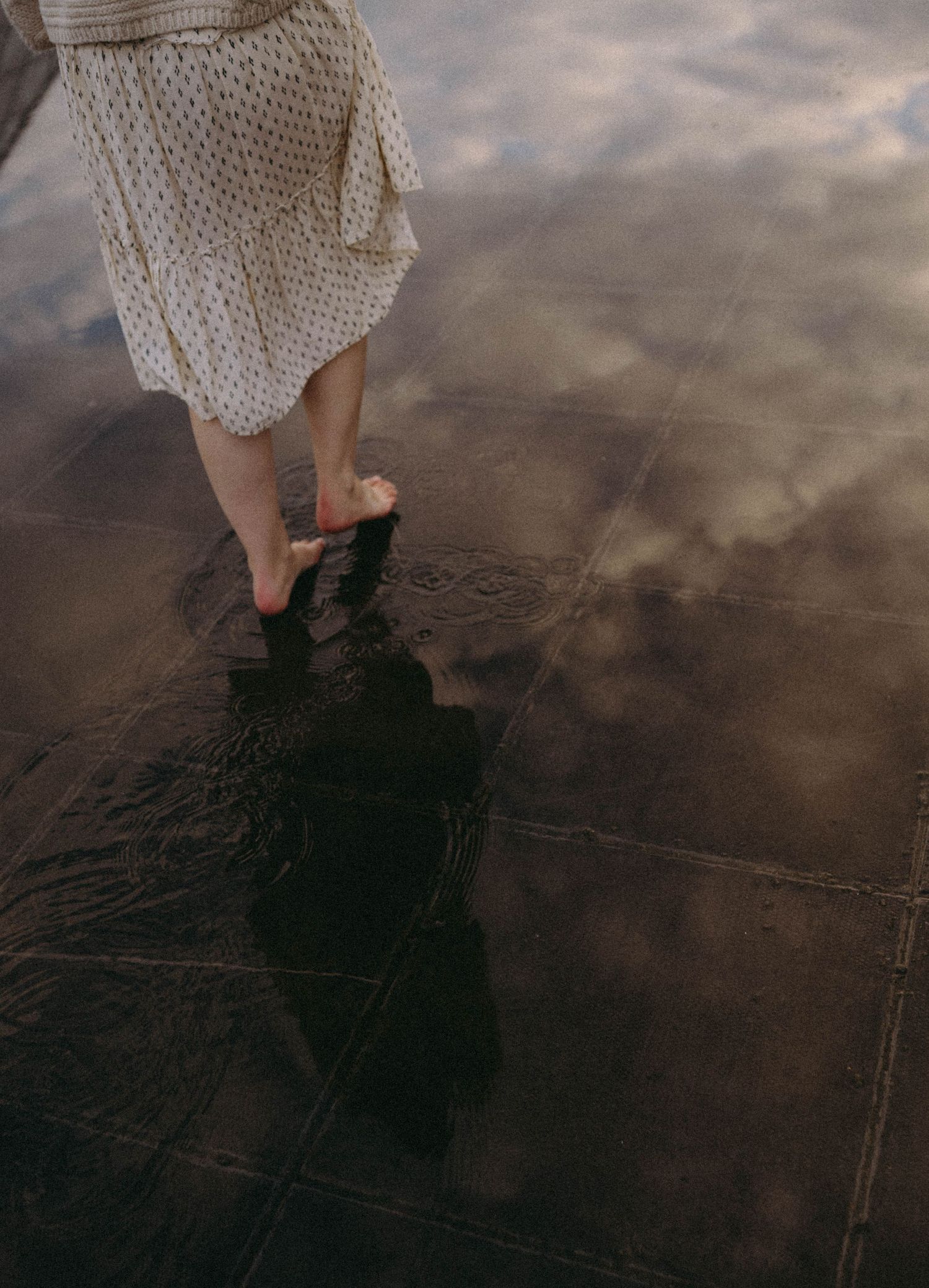 Person wearing white lace dress stands on dark wet sand at sunset, creating a moody coastal atmosphere.