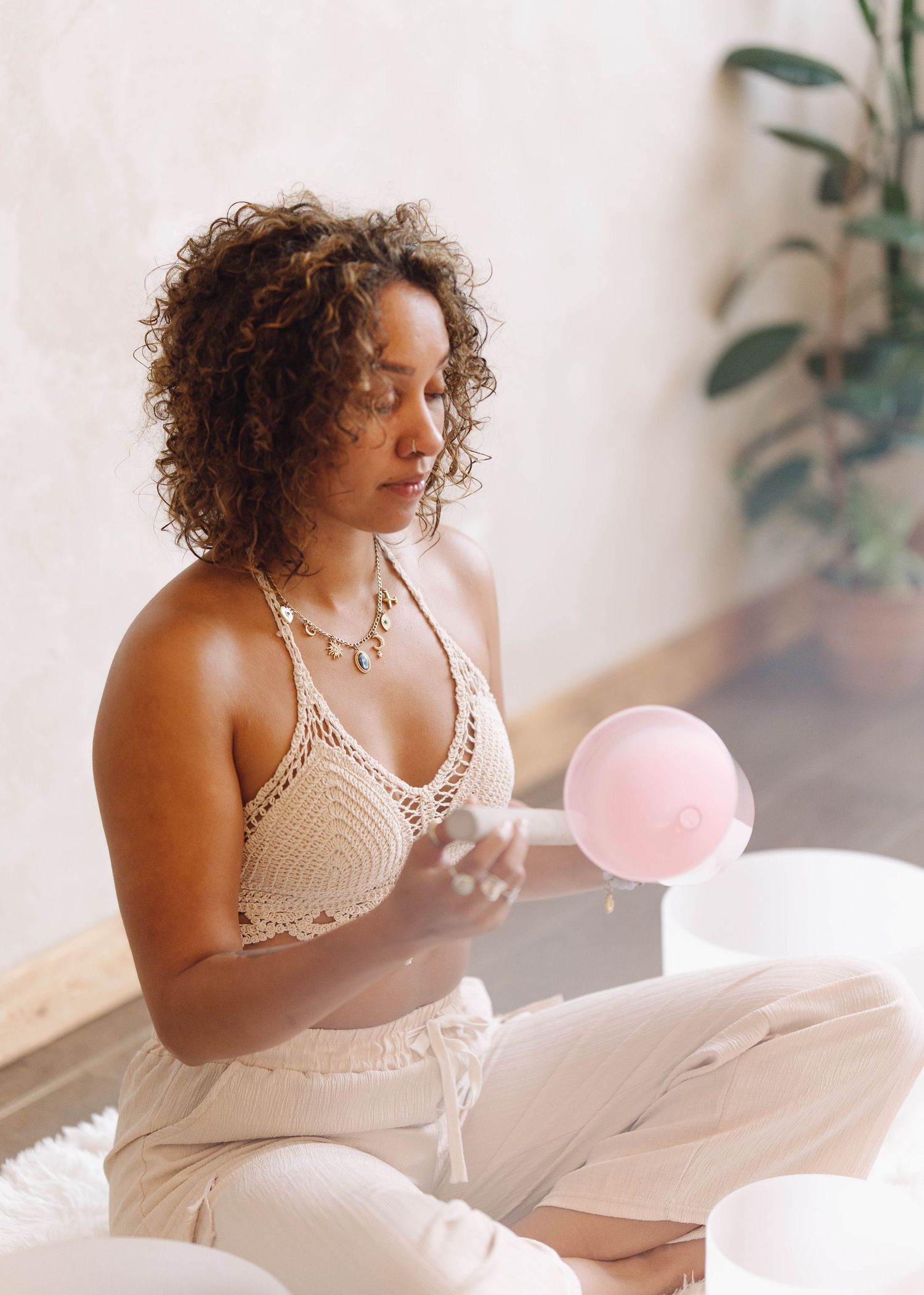 Woman in crochet top playing a pink crystal singing bowl while seated on a white rug indoors.