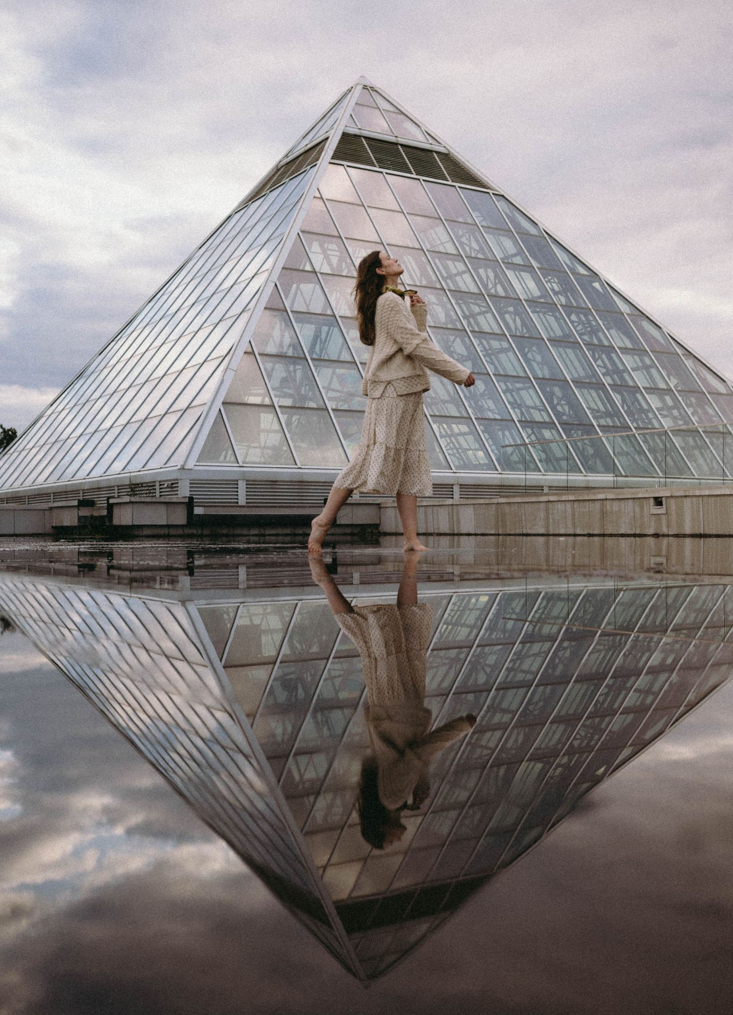 Reflective glass pyramid creates a mirror image on wet ground during overcast weather.