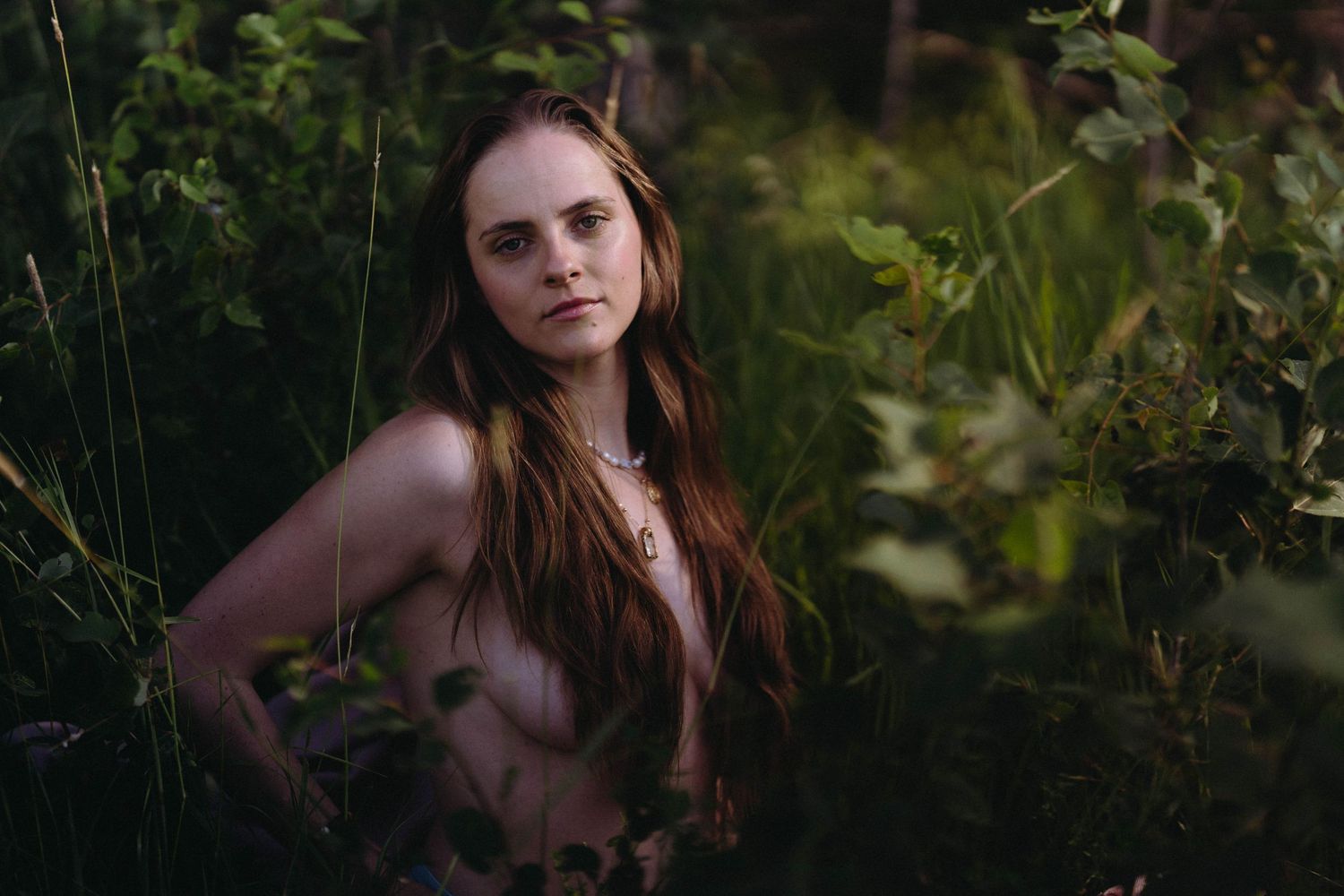 Young woman with long red hair wearing a necklace, surrounded by lush green foliage in a forest setting.