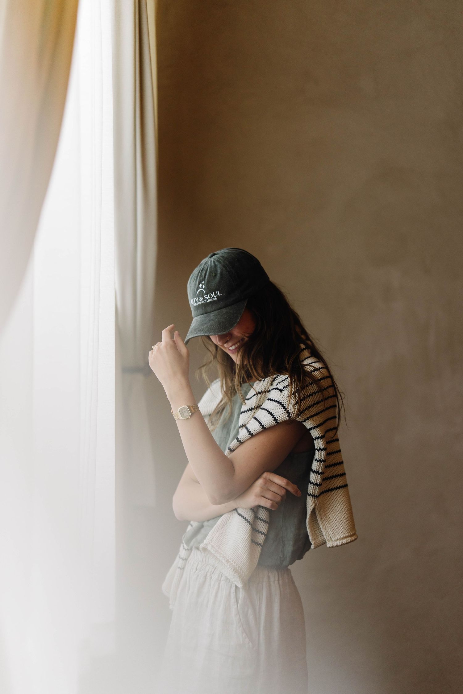 Woman in striped tee, white skirt, green cap, and sunglasses smiling by a curtained window with warm light.