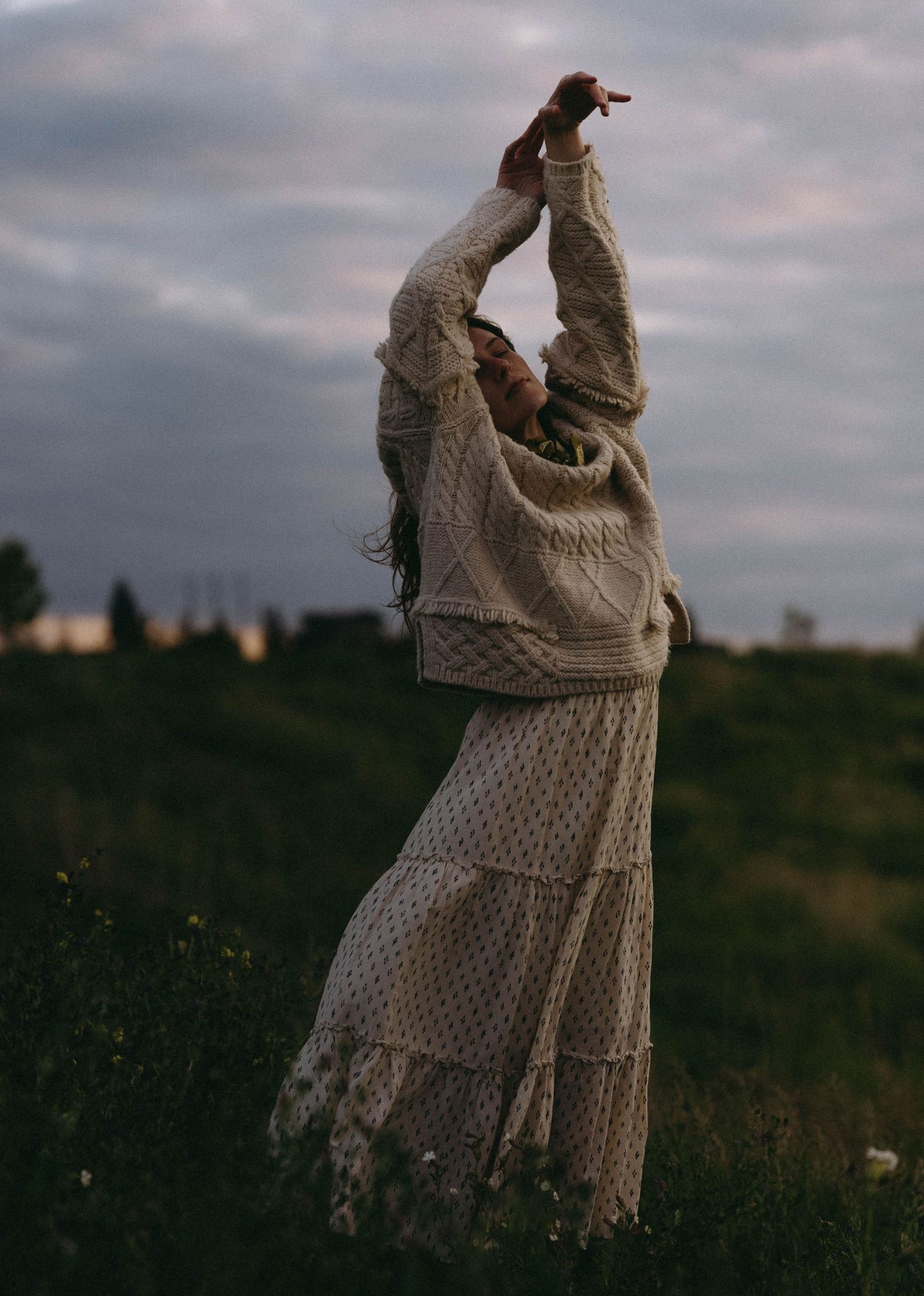 A person in a vintage knit sweater and lace skirt stretches their arms upward against a moody evening sky.