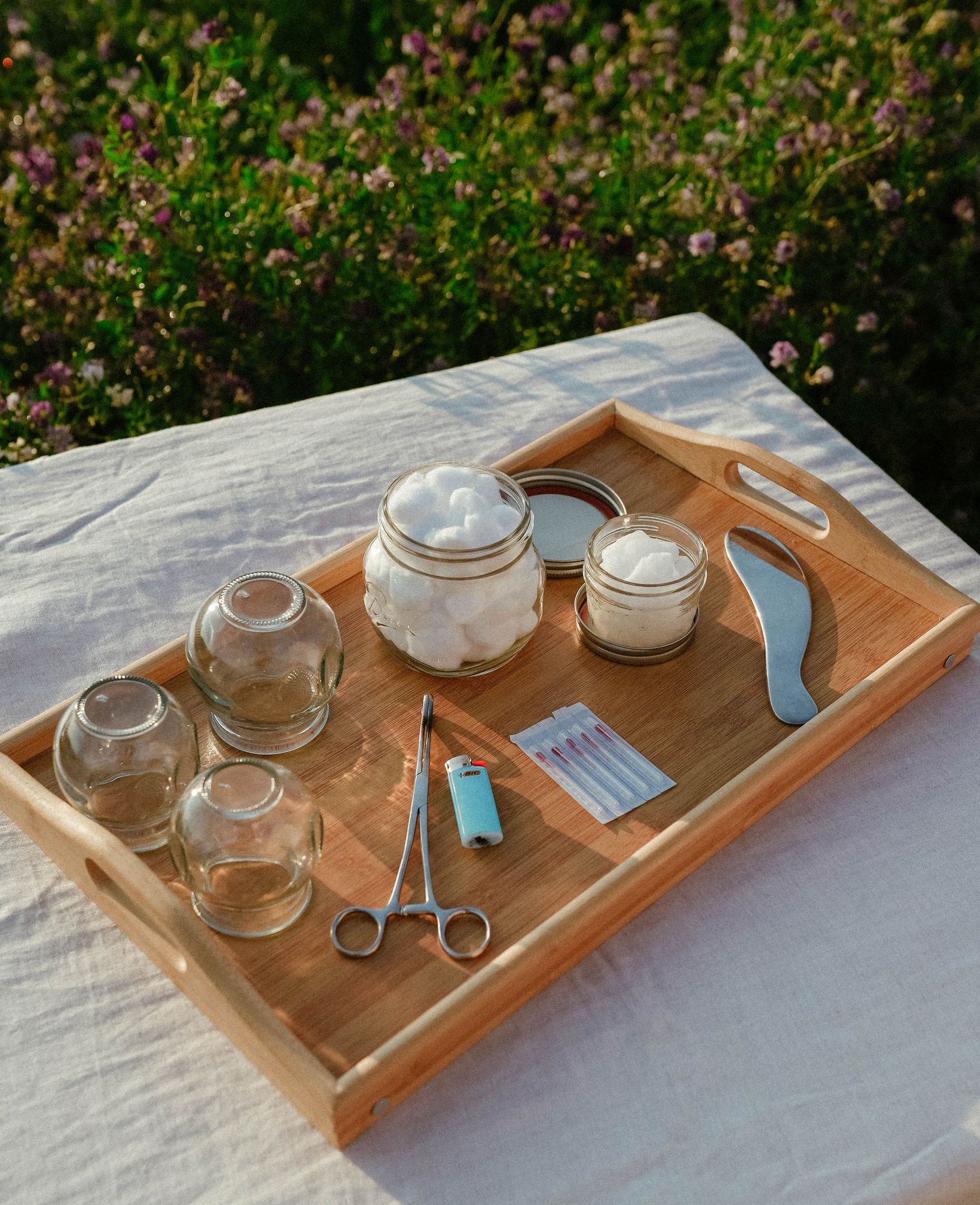 Bamboo tray with glass cupping jars, cotton balls, lighter, forceps, and needles for traditional cupping therapy outdoors.