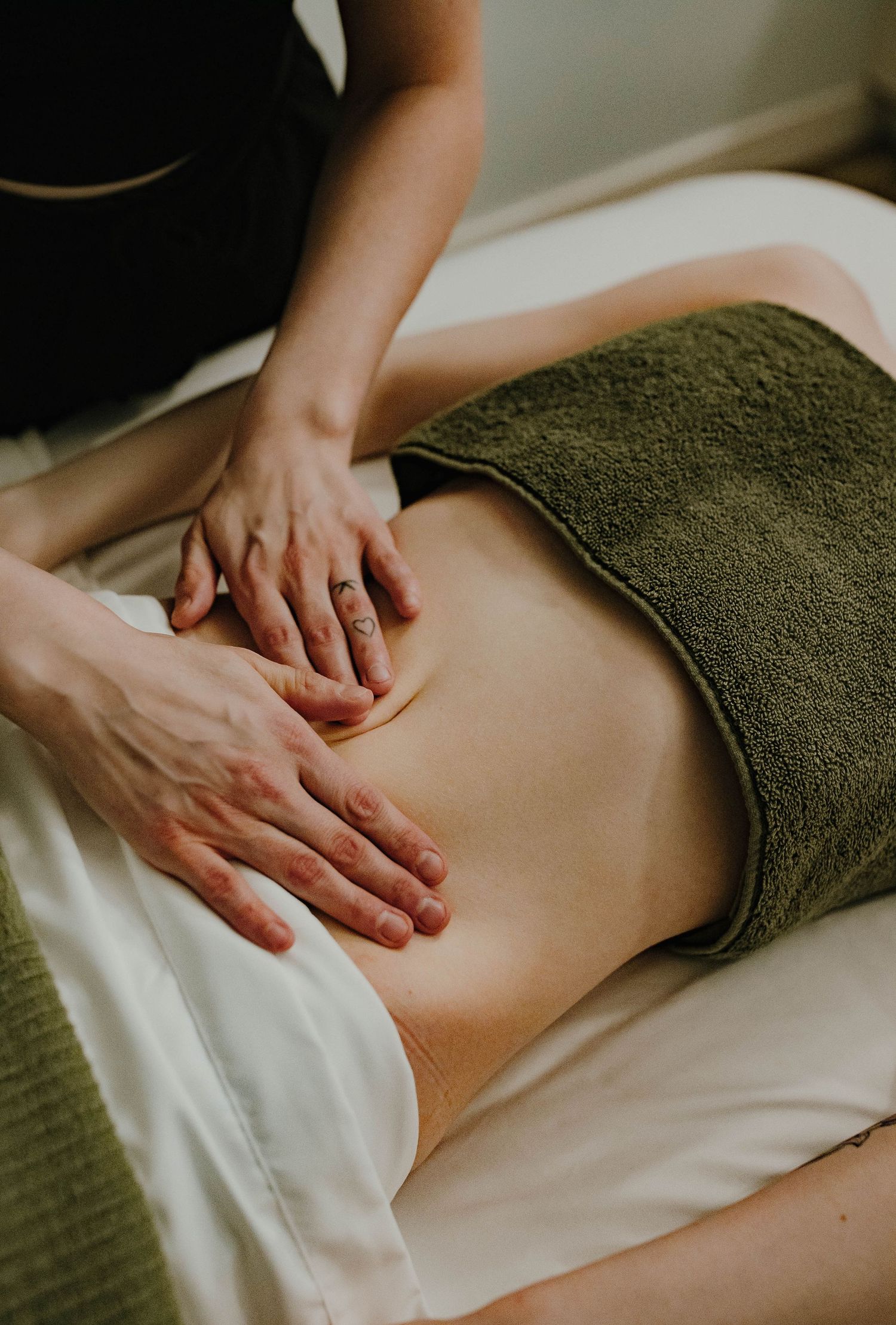 A massage therapist performs an abdominal massage on a client draped with a green towel on a white table.