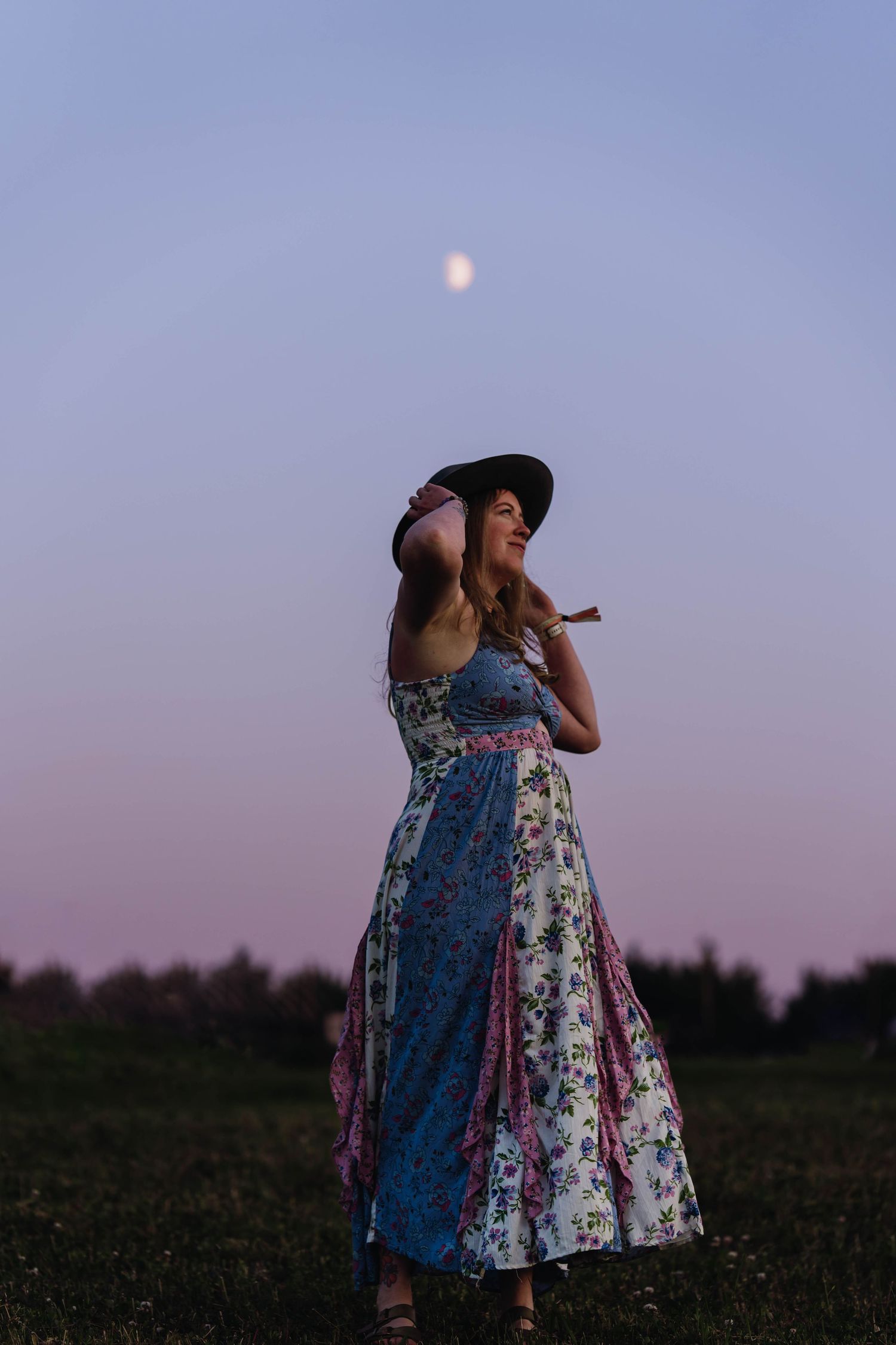 Woman in floral dress and wide-brim hat stands in field at dusk with moon visible in purple sky.