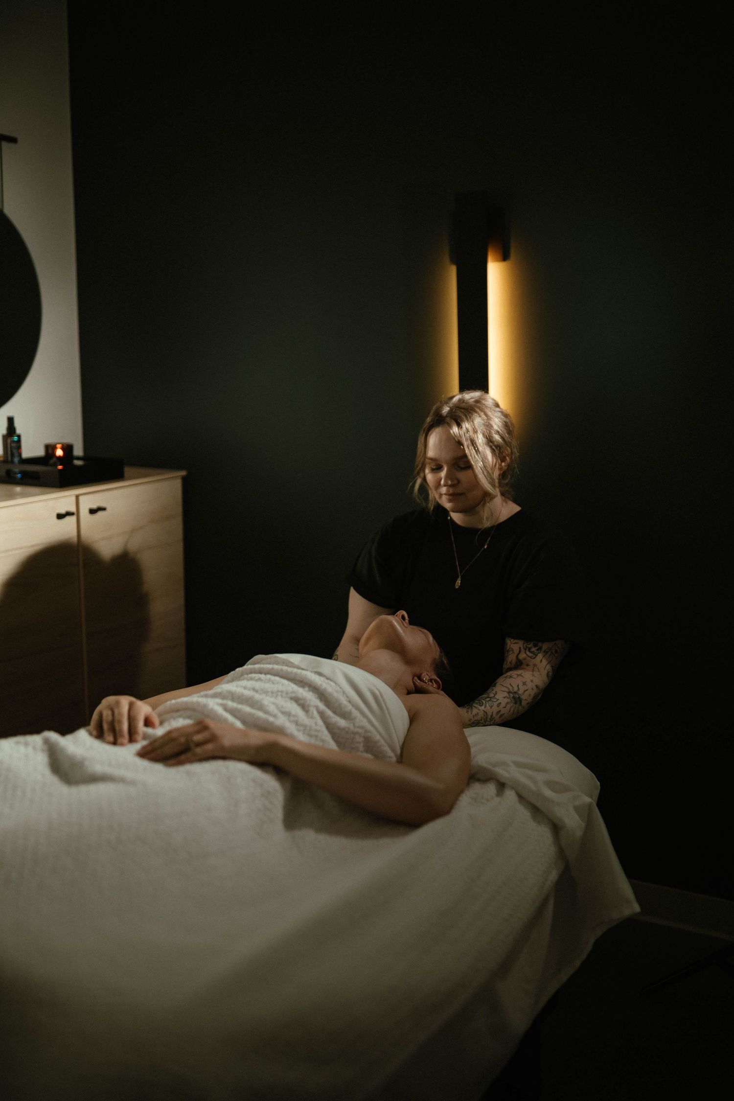 A massage therapist with tattoos performs a relaxing treatment on a client lying on a white draped table in a dimly lit spa.