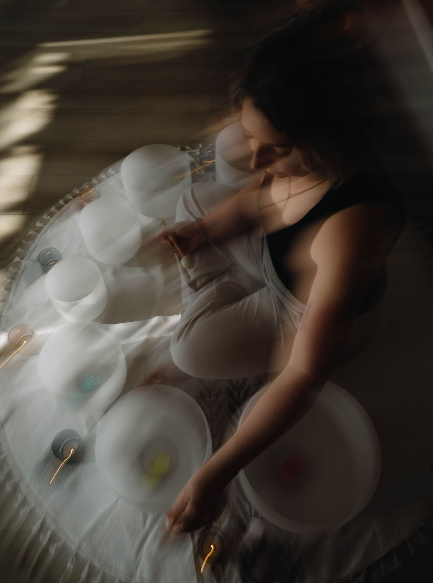 Woman playing crystal singing bowls on the floor, captured with motion blur in soft natural light.