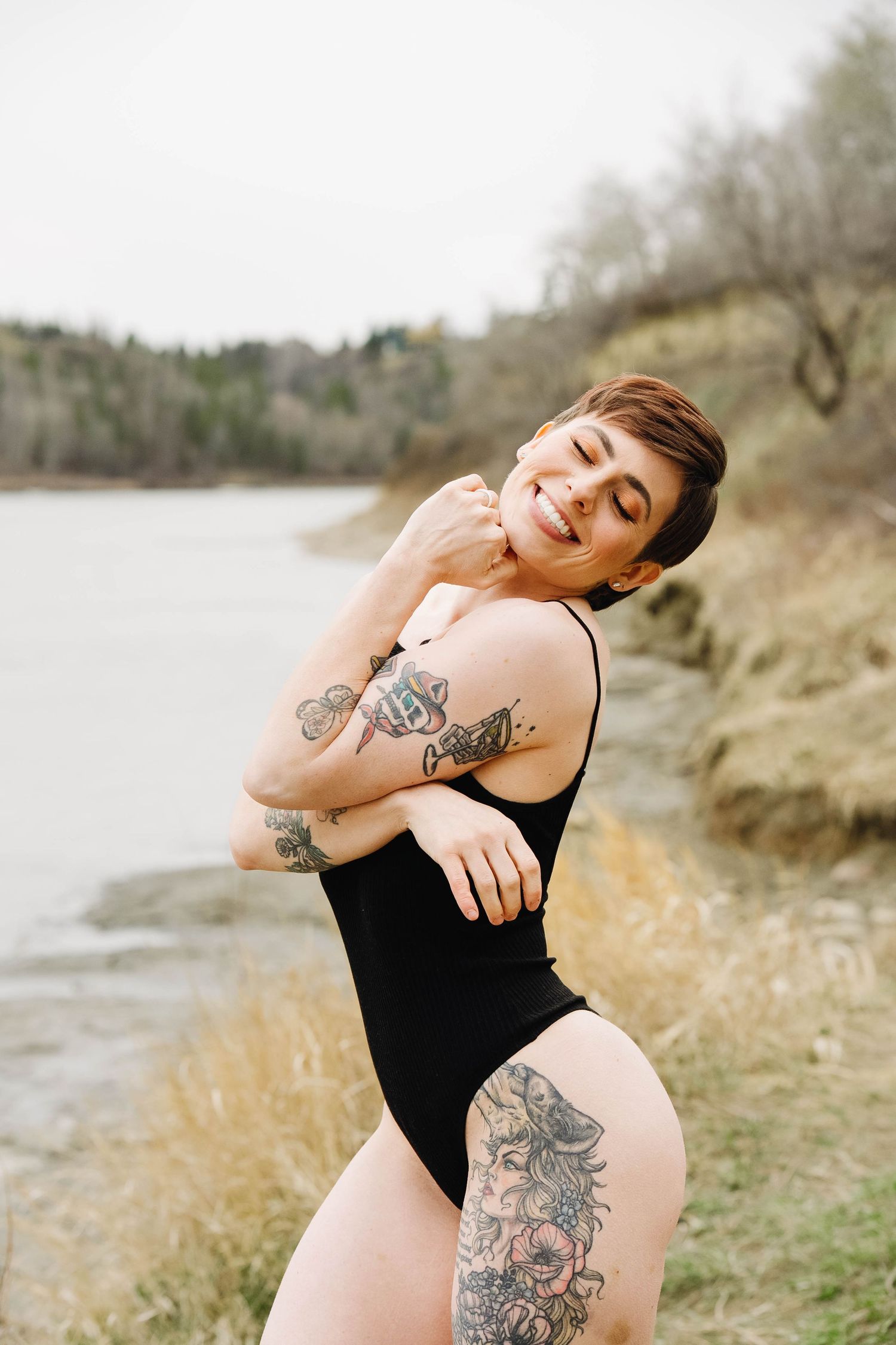 A tattooed woman in a black swimsuit poses near a lake shore with dry grass and trees in the background.