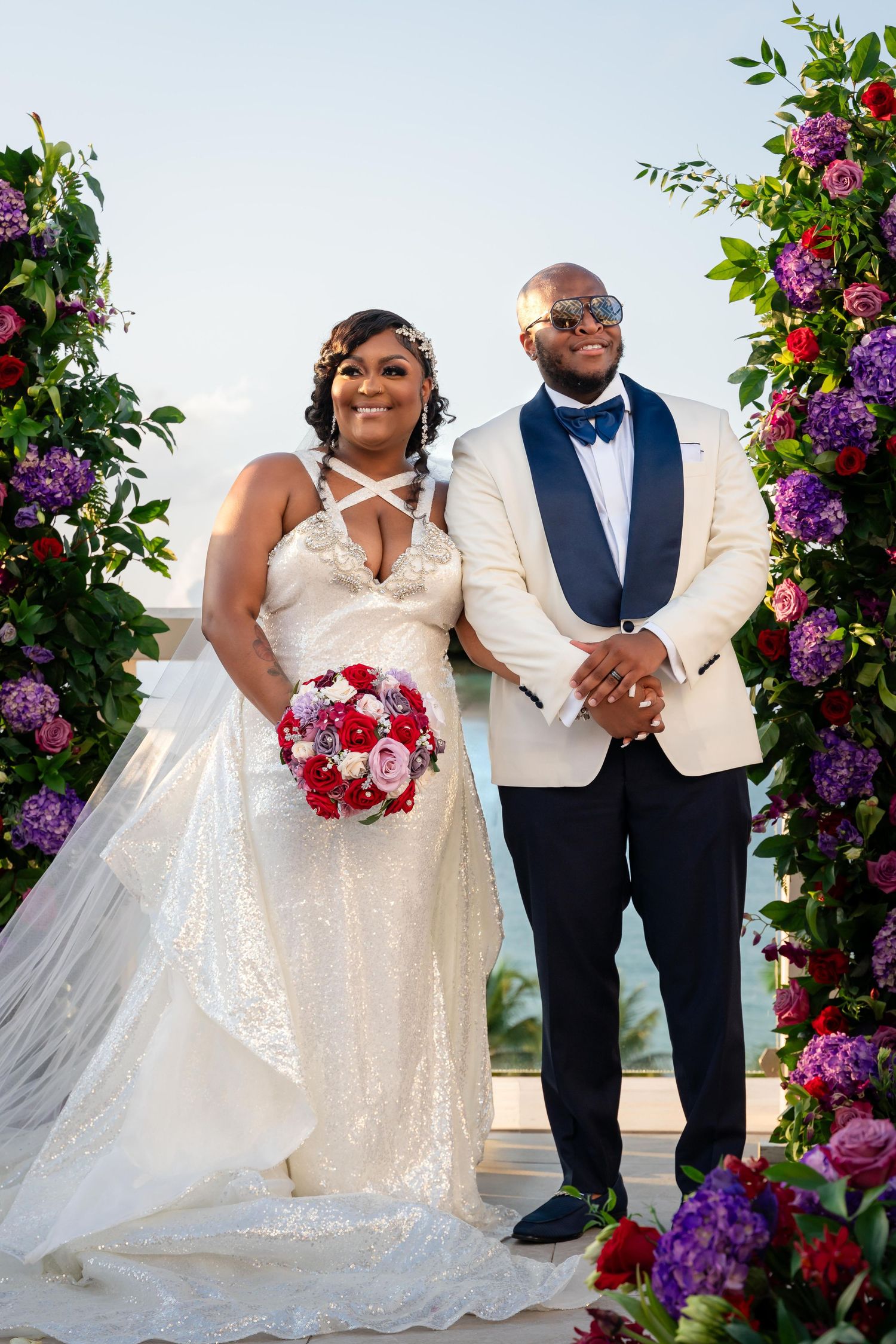 Wedding couple poses under floral arch with purple and pink blooms during outdoor celebration.