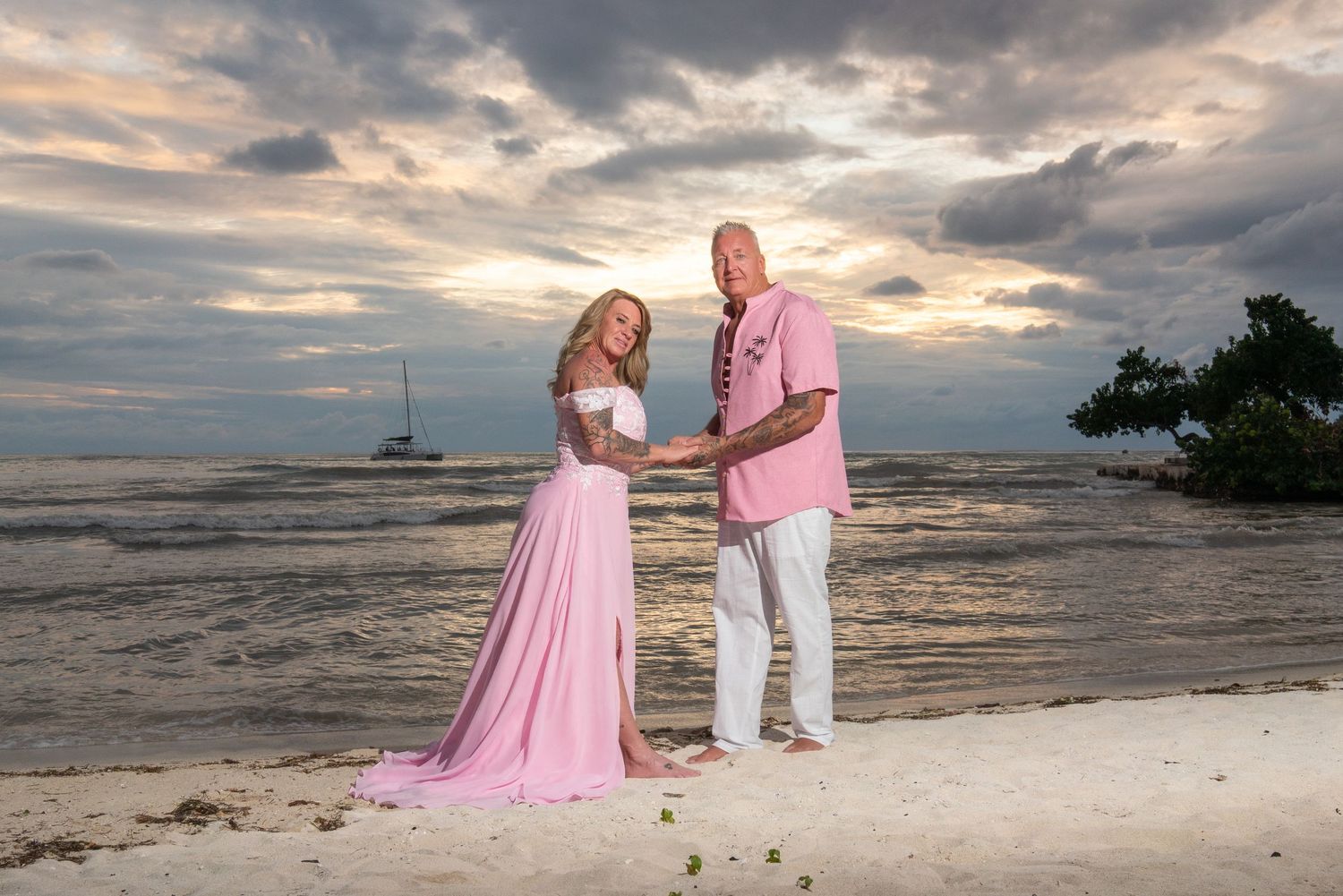 Couple in pink and white outfits posing romantically at sunset on a tropical beach with sailboat in background.