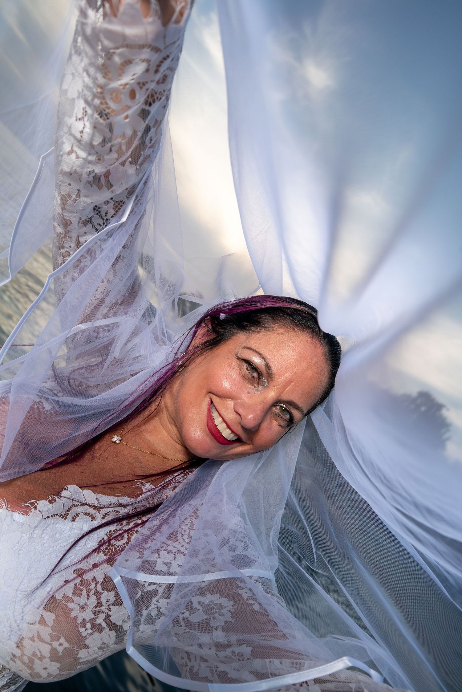 Artistic bridal portrait showing up close of bride's face under flowing white lace veil against a blue sky background.