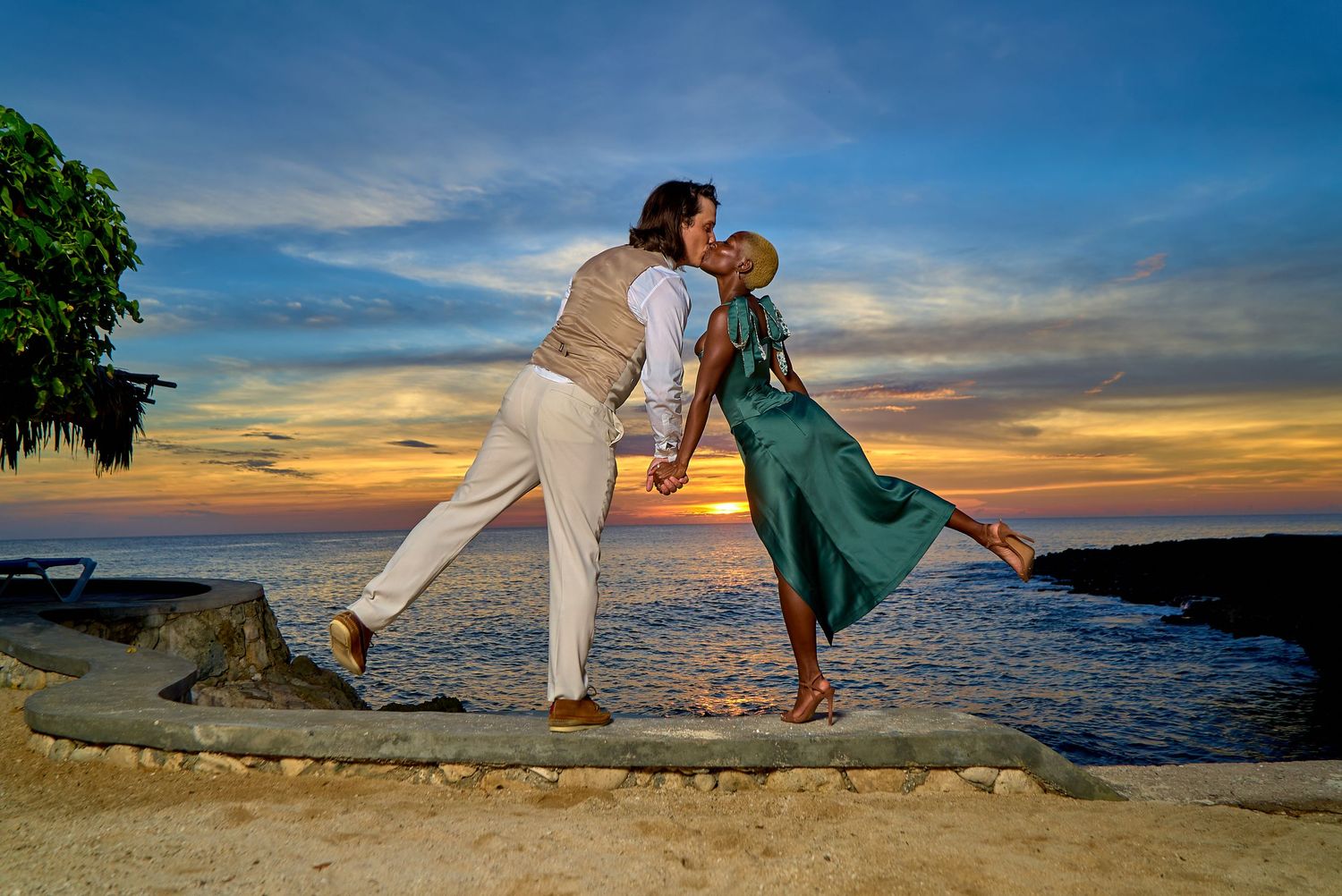 A romantic beachside photo at sunset with two people striking a playful pose on the rocky shoreline.