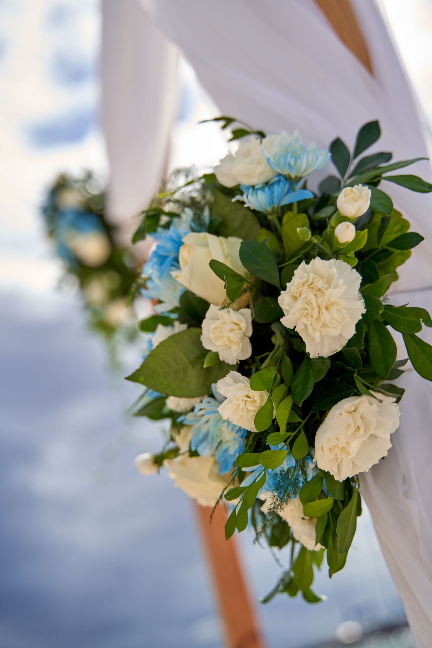 White carnations and blue flowers decorate a white wedding arch against a blurred outdoor background.