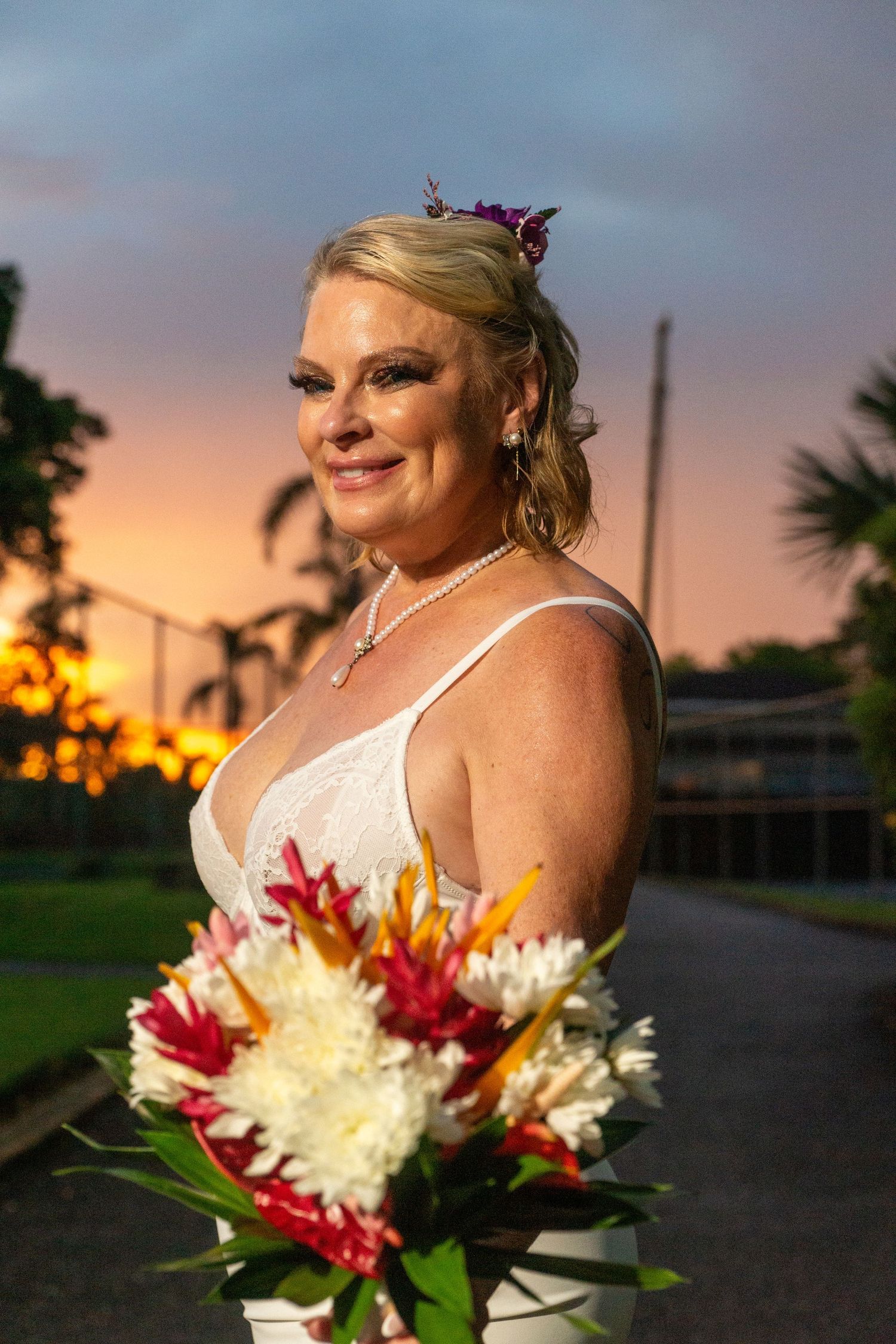 Bride holds colorful bouquet of red and white flowers against sunset sky with palm trees in background.