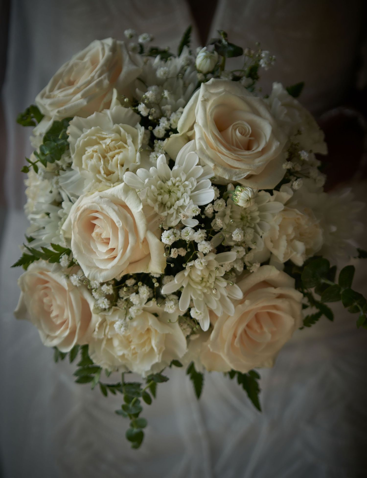 An elegant white wedding bouquet featuring cream roses and baby's breath arranged with delicate greenery.
