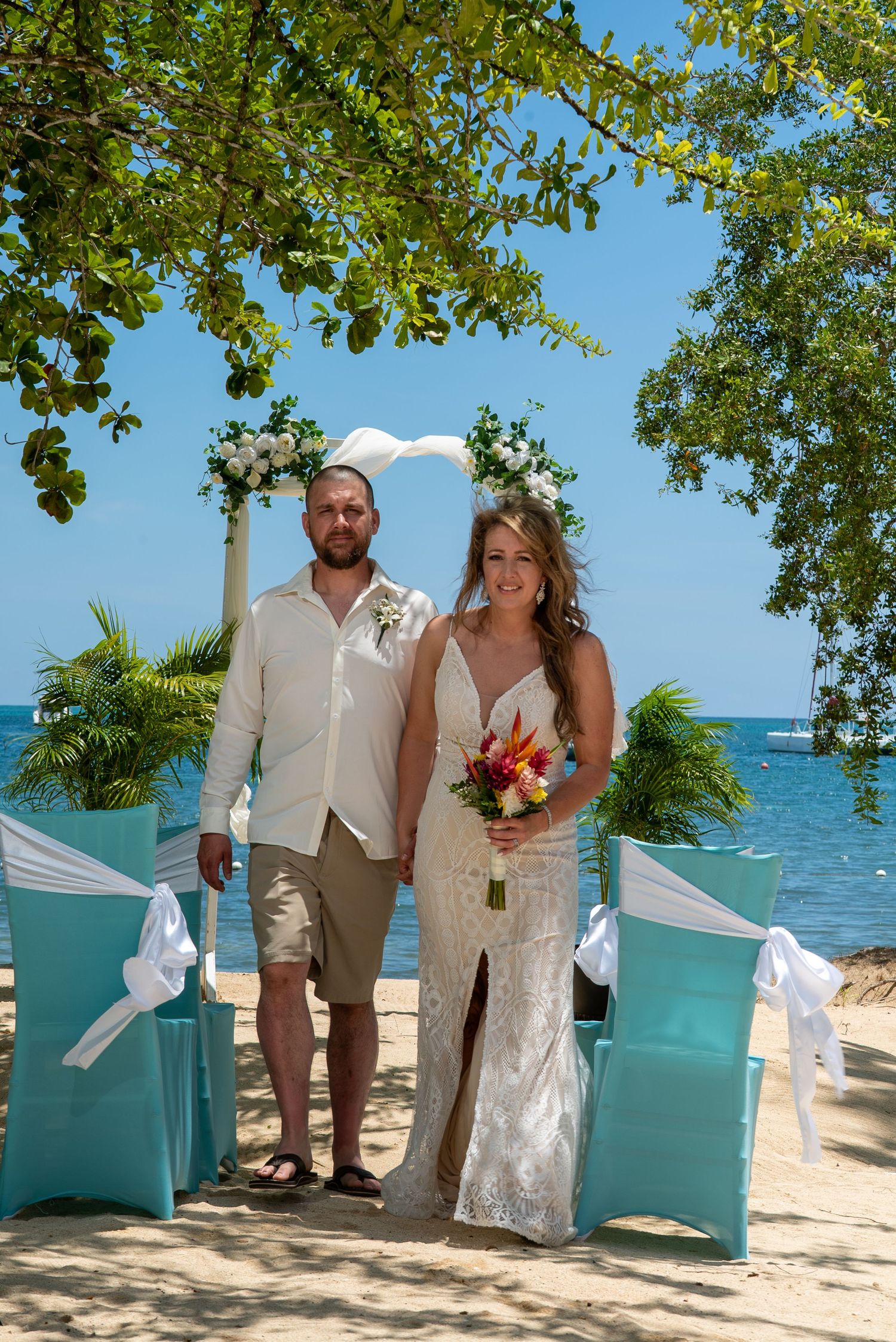 Romantic beach wedding ceremony scene with turquoise chairs and tropical ocean backdrop under green trees.