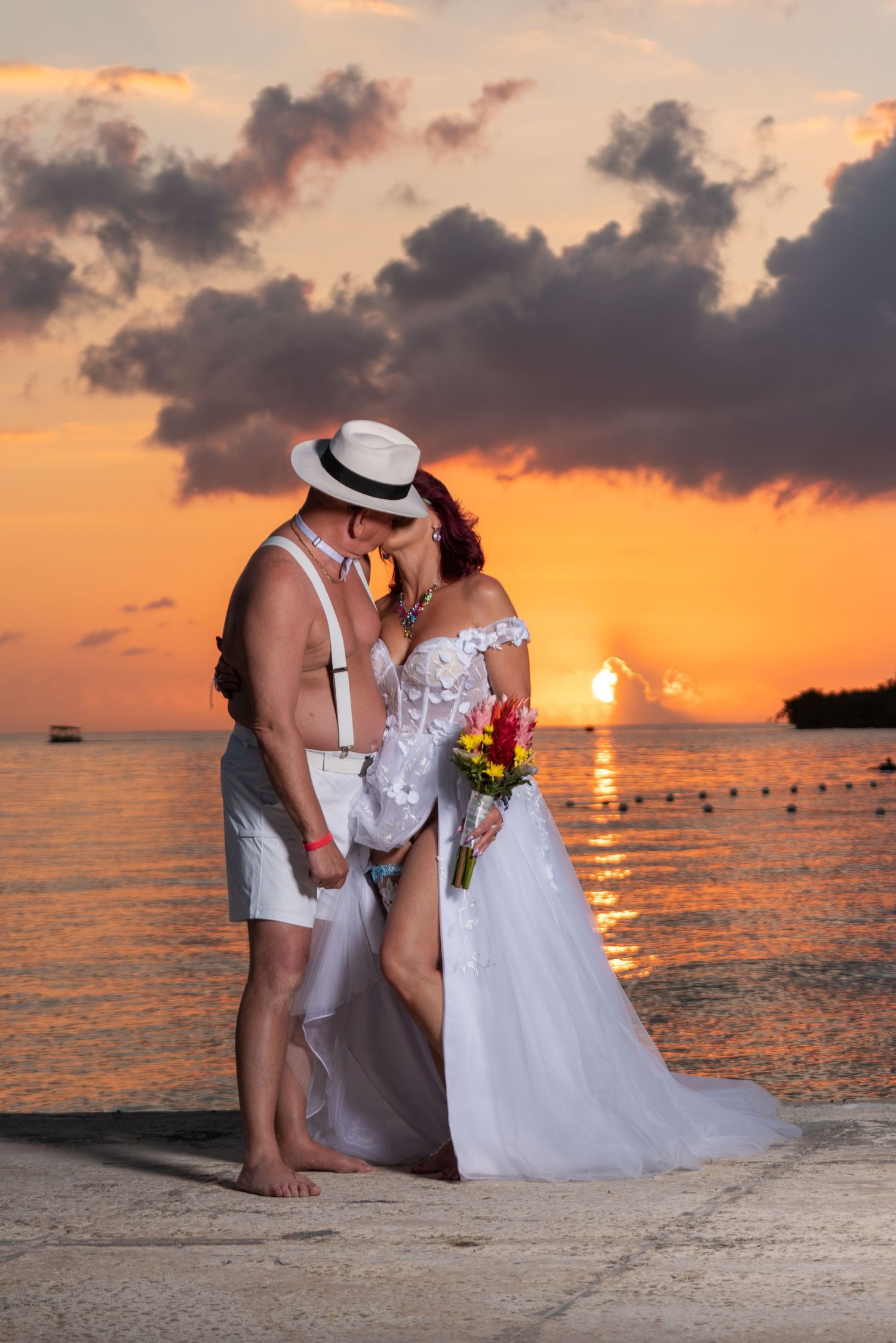 Couple in wedding attire shares romantic moment on beach at sunset with orange sky and dark clouds over ocean.