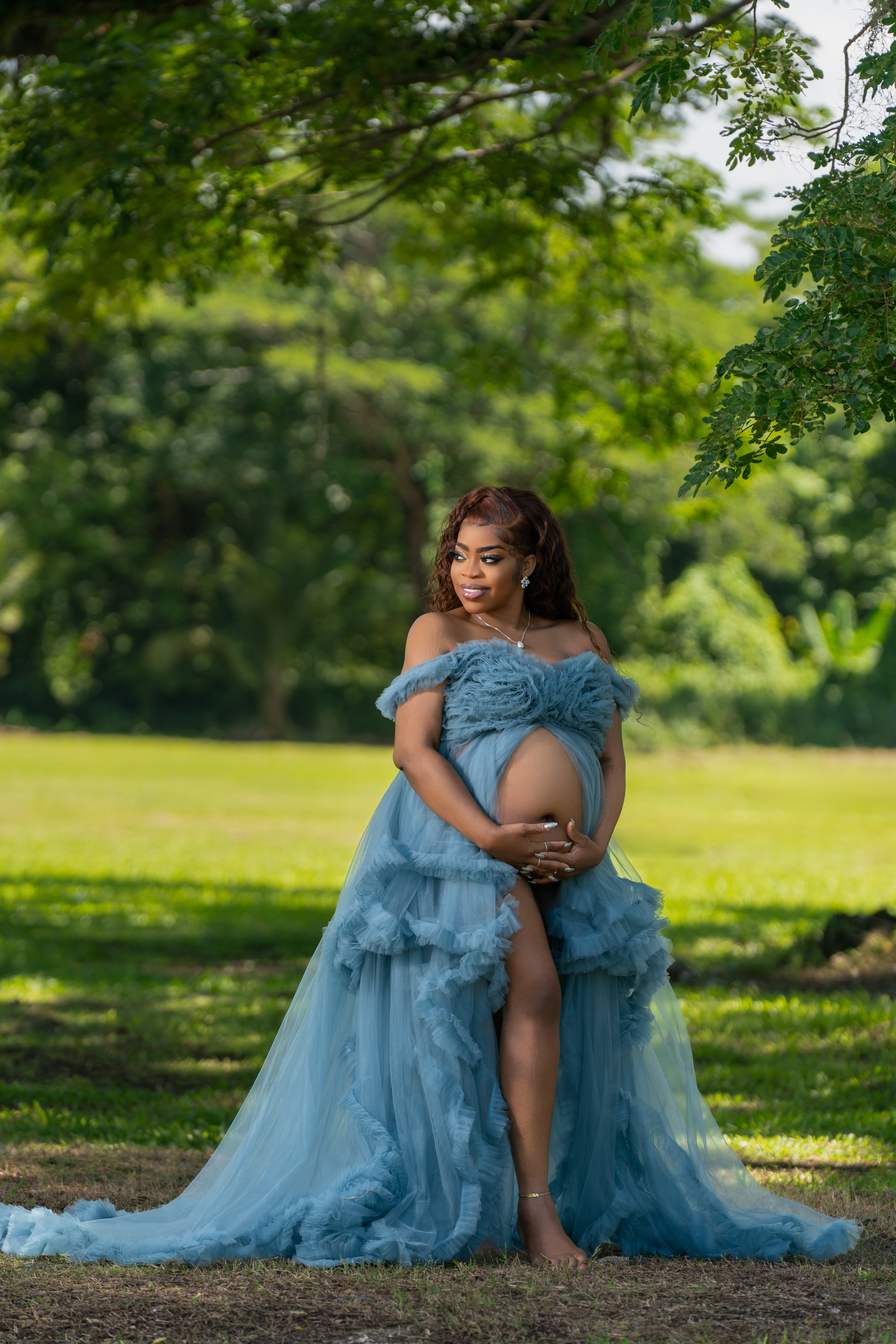 Ethereal maternity photo shoot in blue tulle dress outdoors under lush green trees.