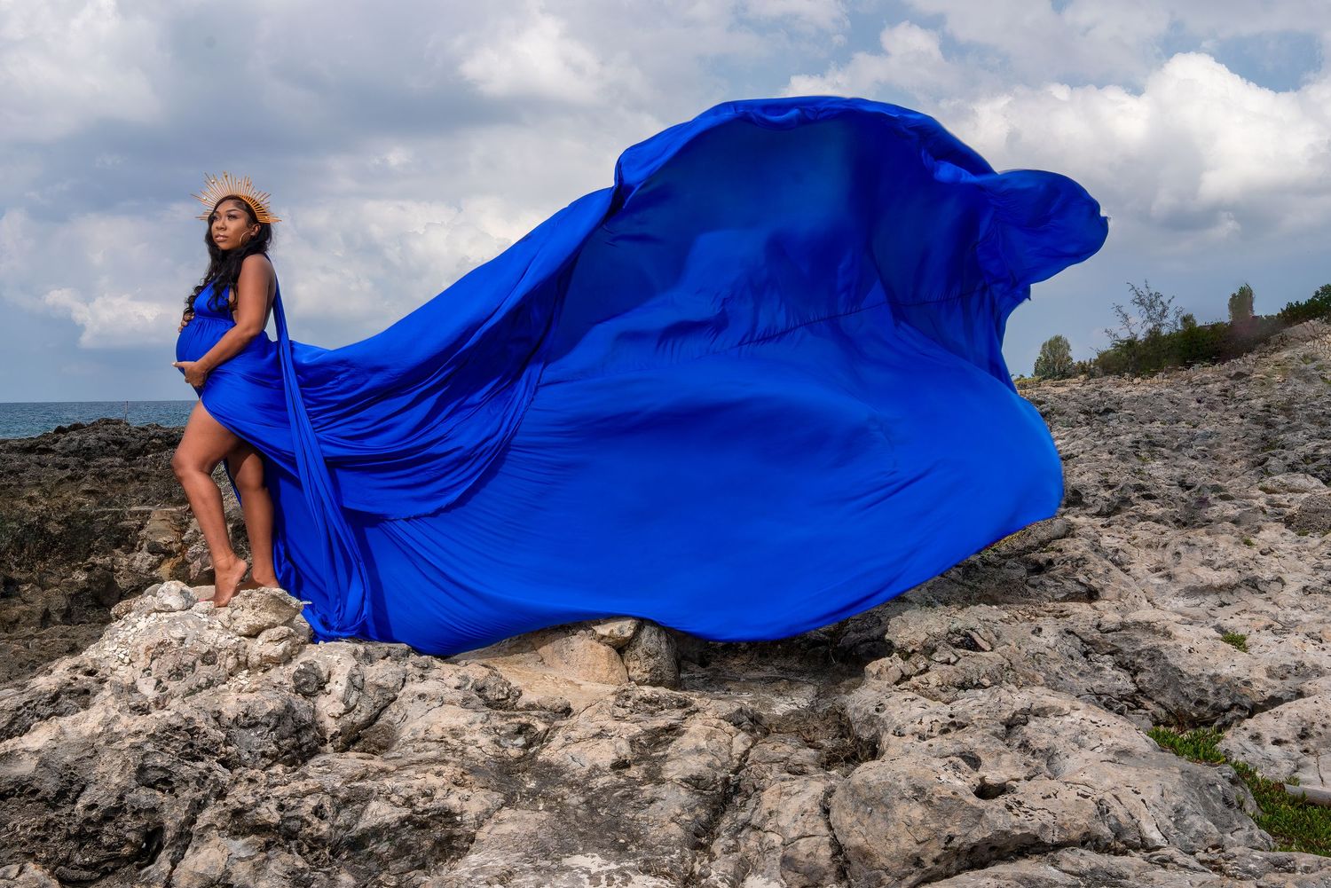 A pregnant woman in a flying blue dress poses on rocky cliffs for a maternity photo shoot.