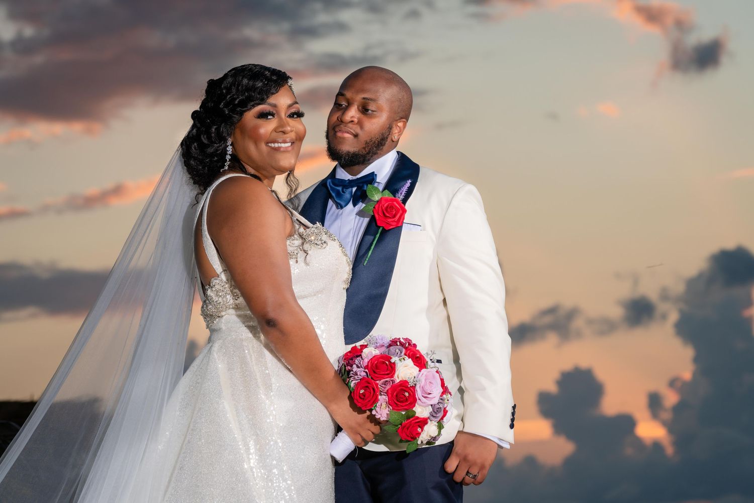 Wedding couple embraces against dramatic sunset sky on beach with pink and red floral bouquet details.