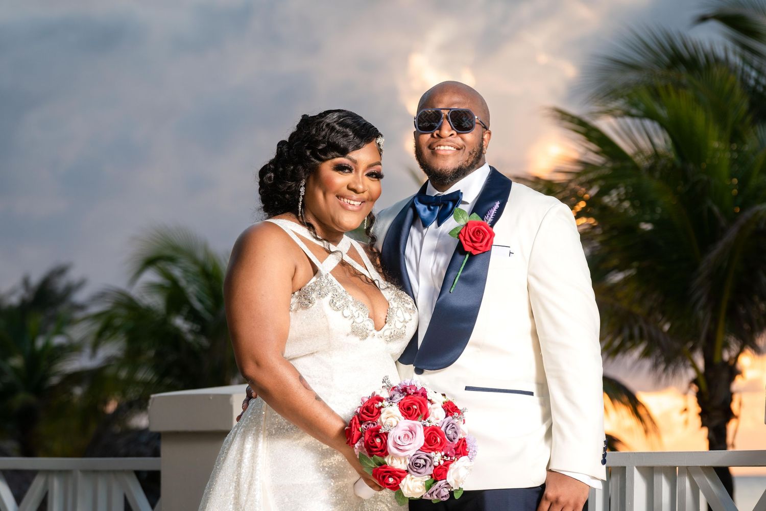 A couple poses together during sunset at a tropical wedding venue with palm trees and red floral bouquet.