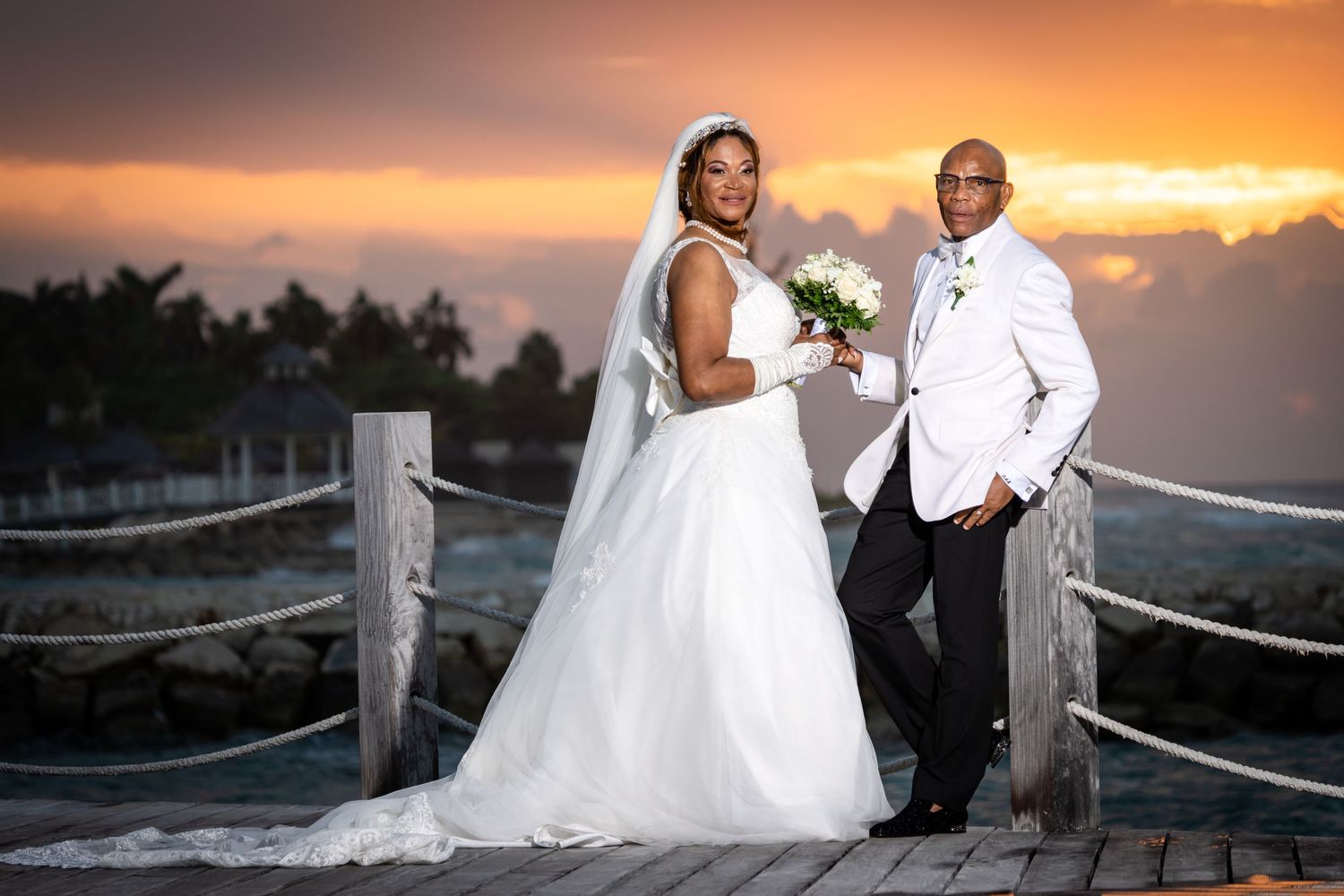 A couple poses on a beach boardwalk during sunset while wearing formal attire and wedding dress.