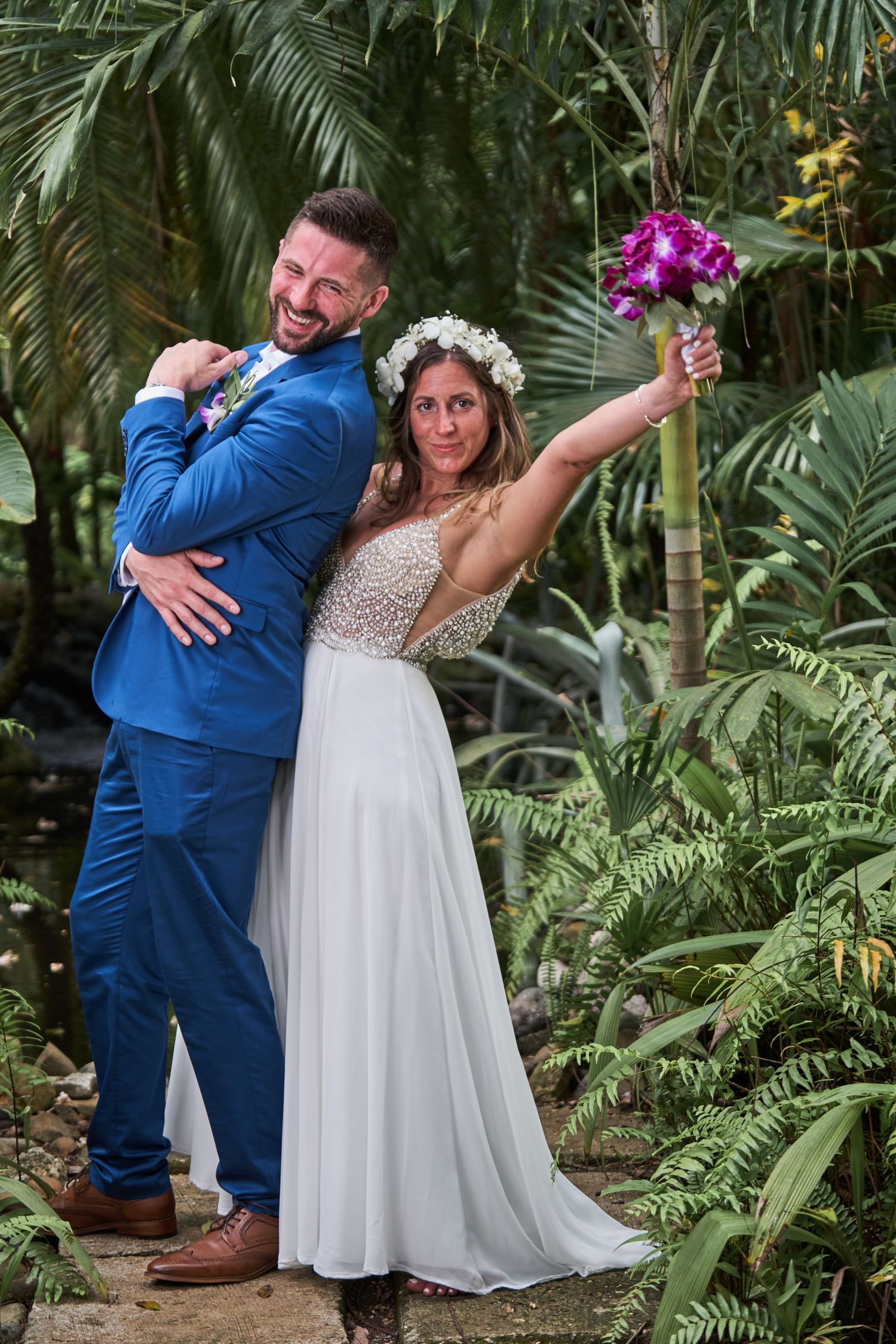 A couple dances joyfully among lush tropical palm fronds at an outdoor garden wedding ceremony.