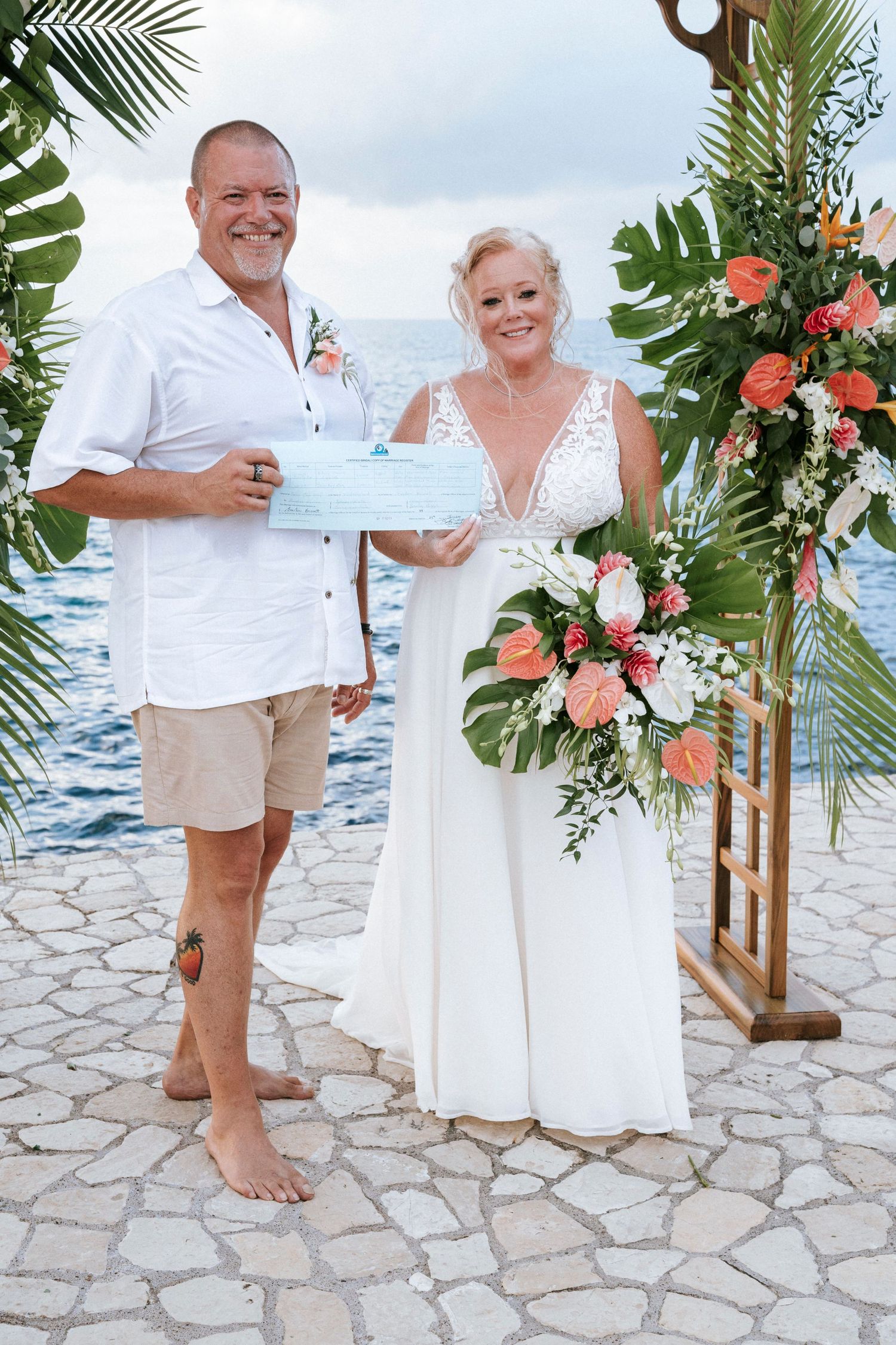 A couple shows their marriage certificate during their tropical beach wedding on a stone walkway with floral decor.