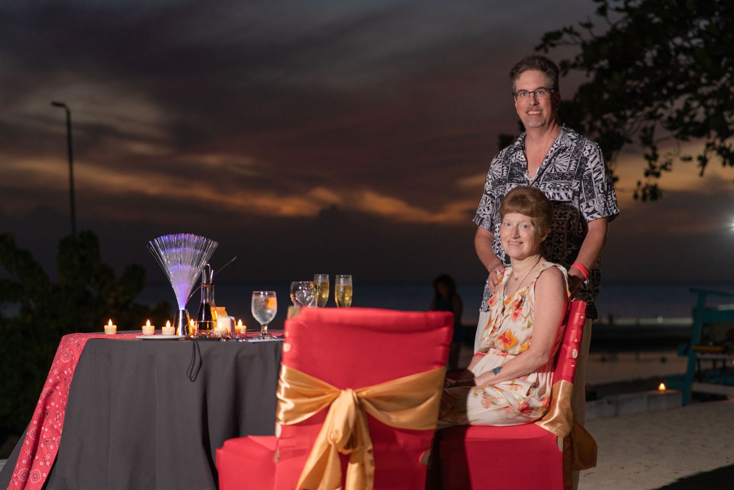 Beachside private dinner setup with red tablecloths and gold accents at sunset showing multiple tables on the sand.