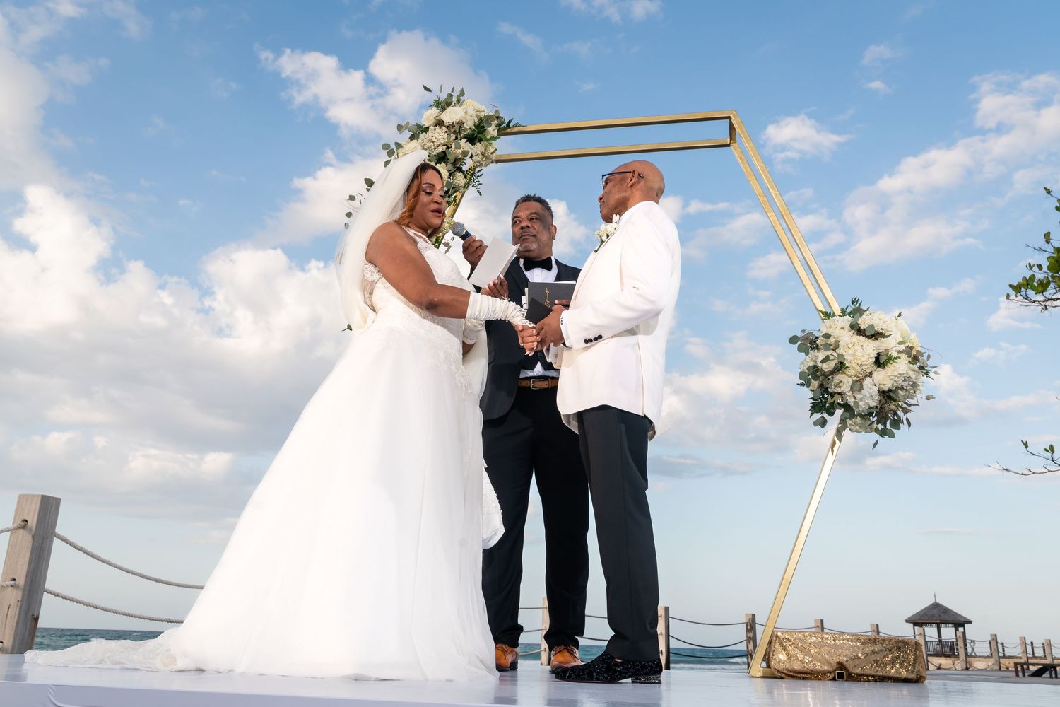 Sunny outdoor wedding ceremony with a gold arch and white floral arrangements against a blue sky.