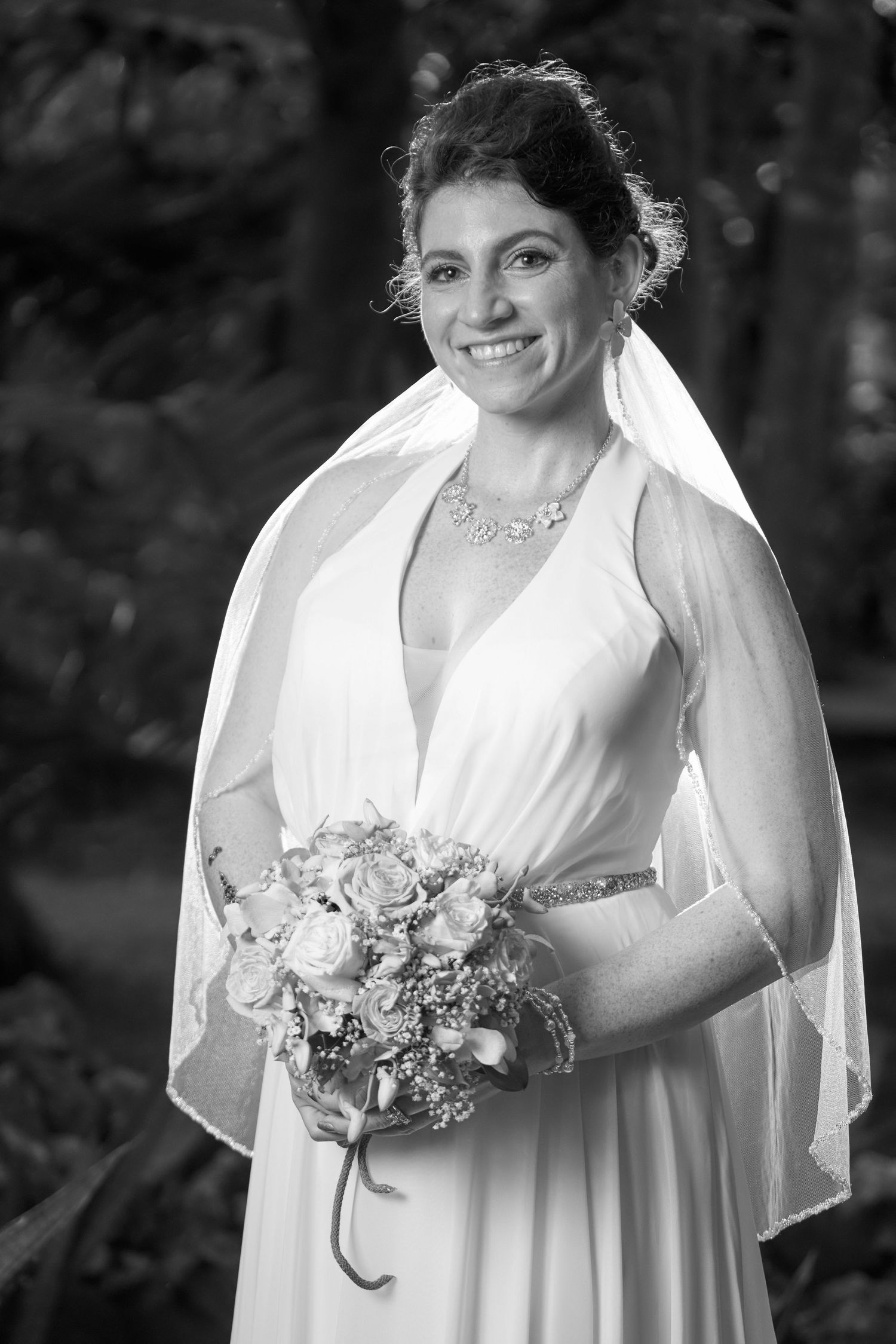 A bride in a flowing white wedding dress carries a delicate bouquet of flowers while posing outdoors in black and white.