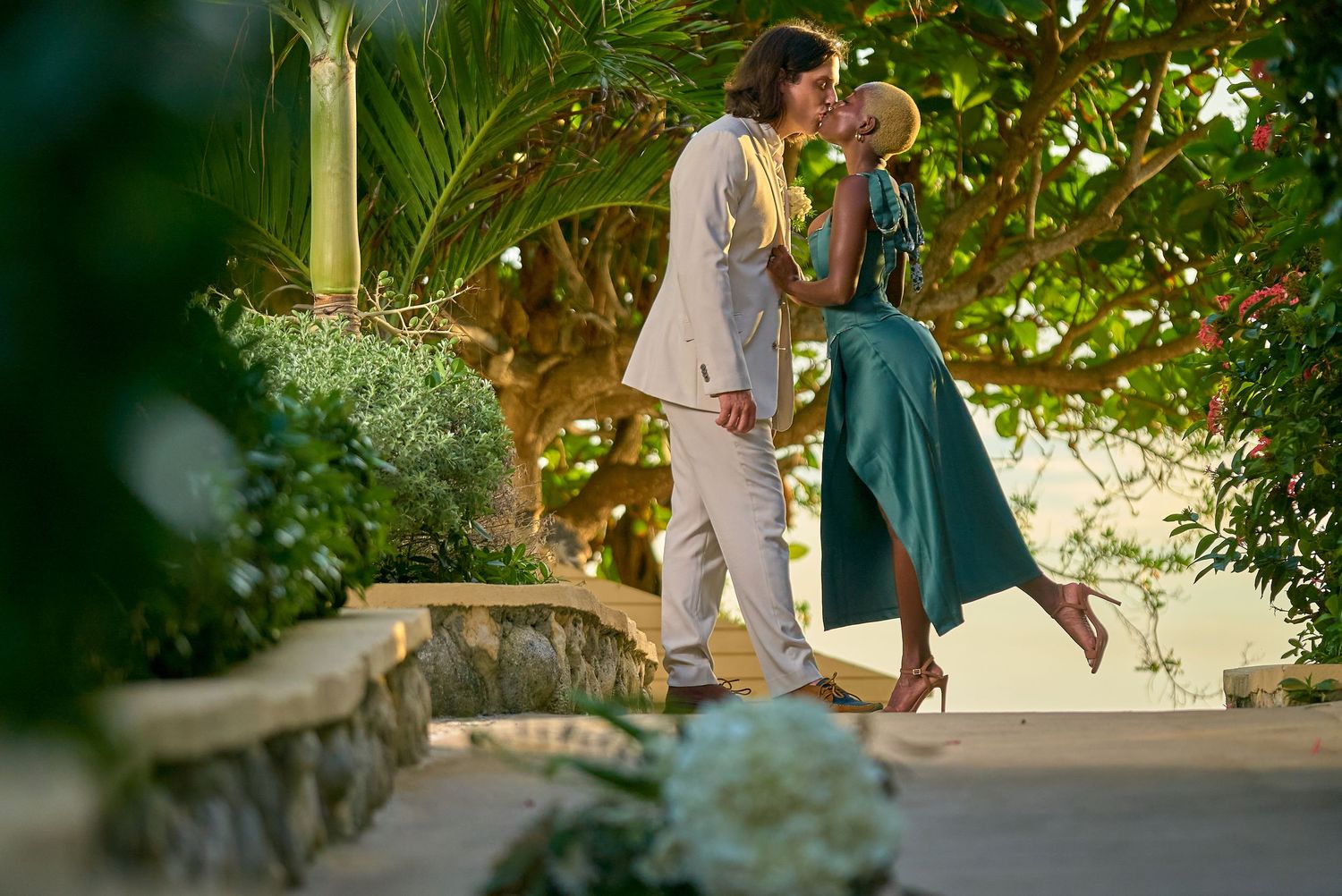 A romantic couple wearing elegant attire strolls amidst lush wedding venue greenery and decorative lights.