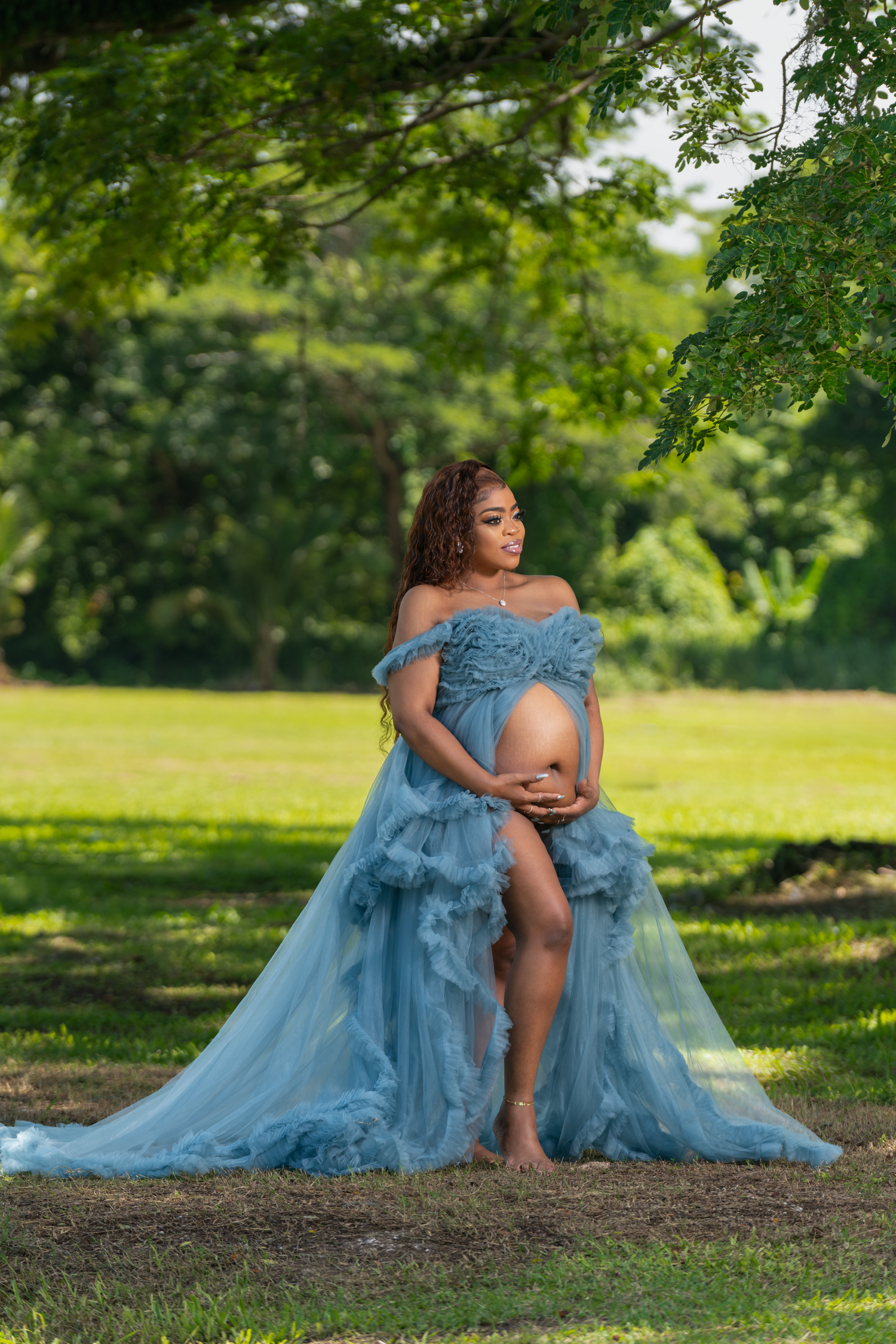 An ethereal figure in a flowing dusty blue tulle gown sits gracefully beneath green trees in a sunlit park setting.