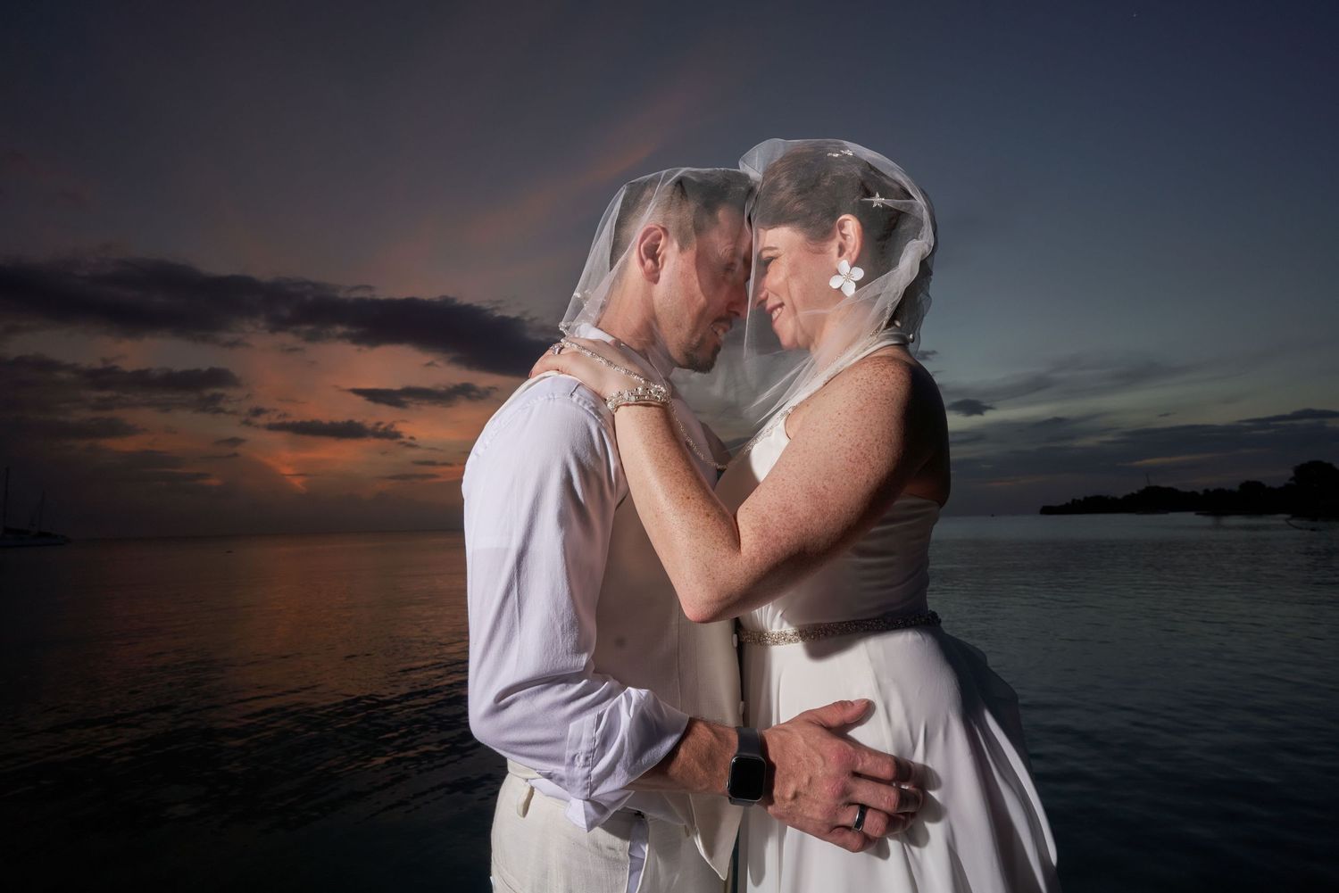 Romantic couple embraces during sunset by the ocean with dramatic sky in background.