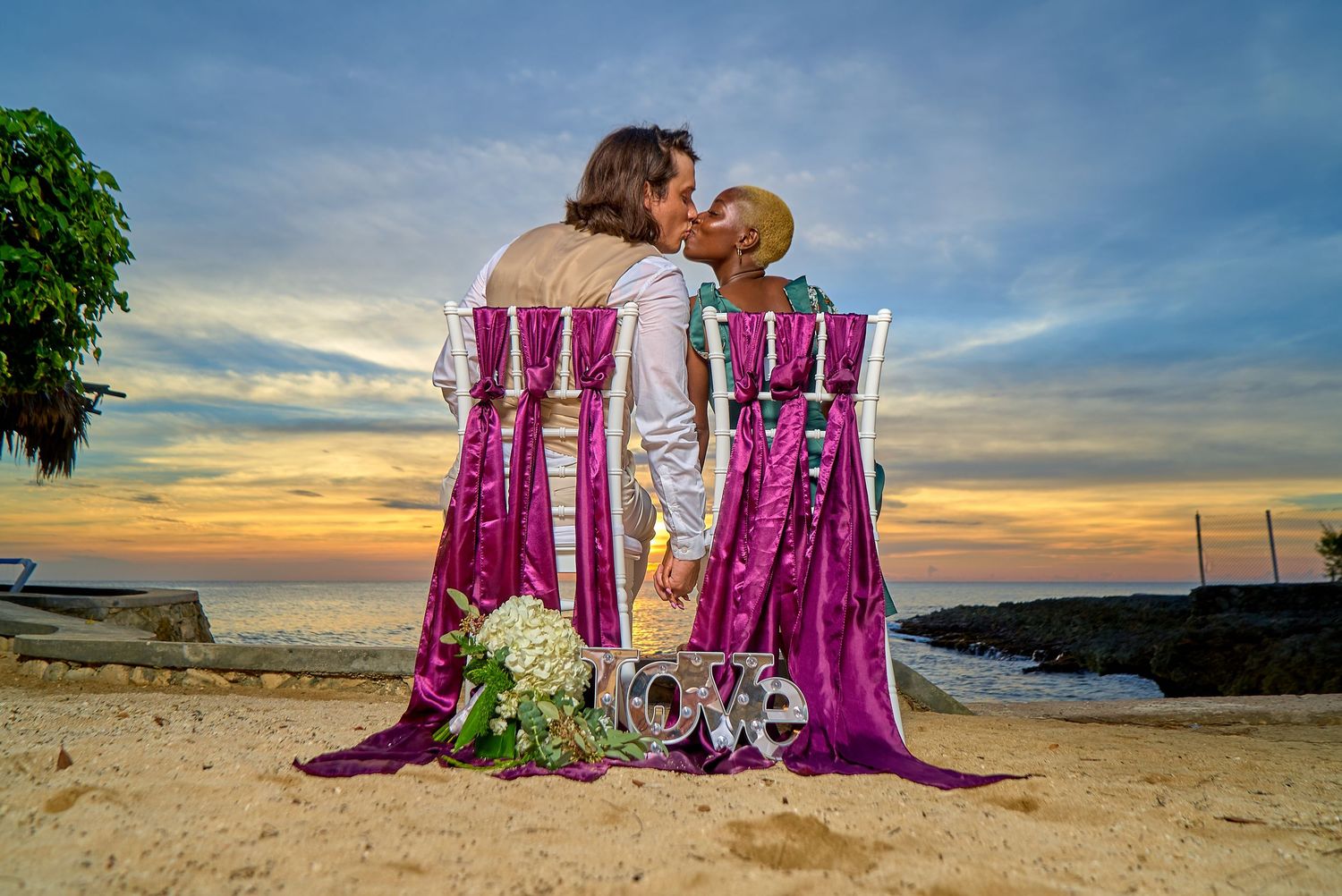 Two people sharing a romantic moment on a beach at sunset with pink chairs and a 'love' sign.