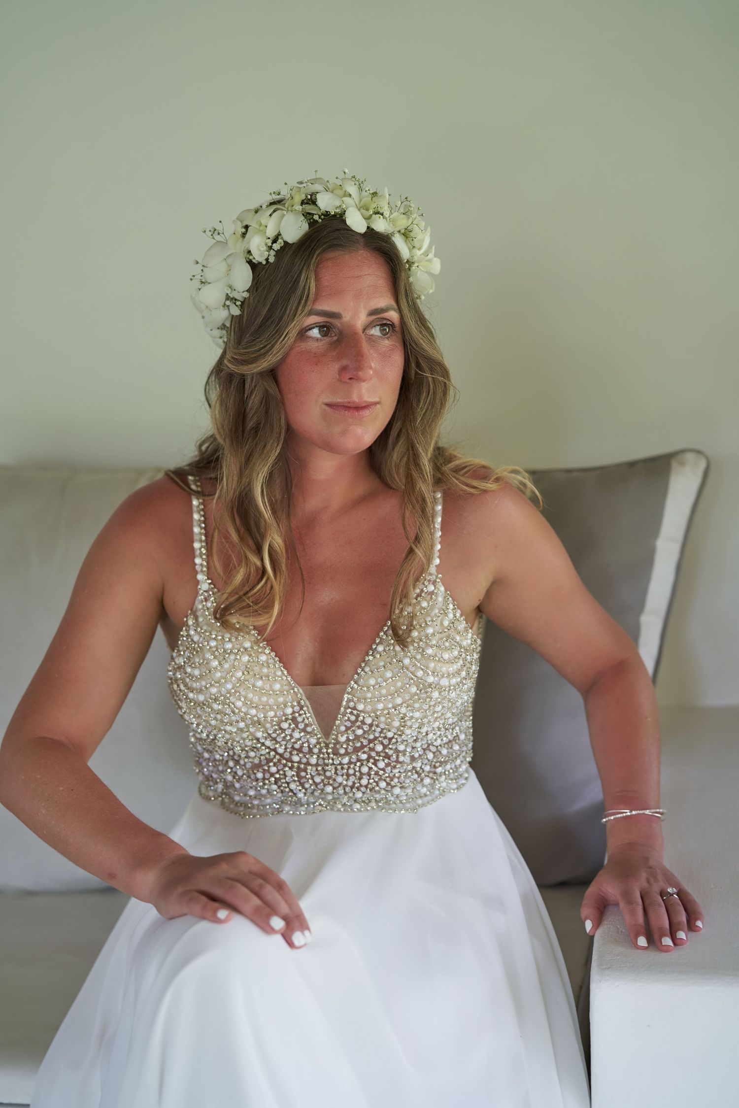 Bride sits on a sofa wearing white tulle wedding dress with beaded bodice and floral crown headpiece.