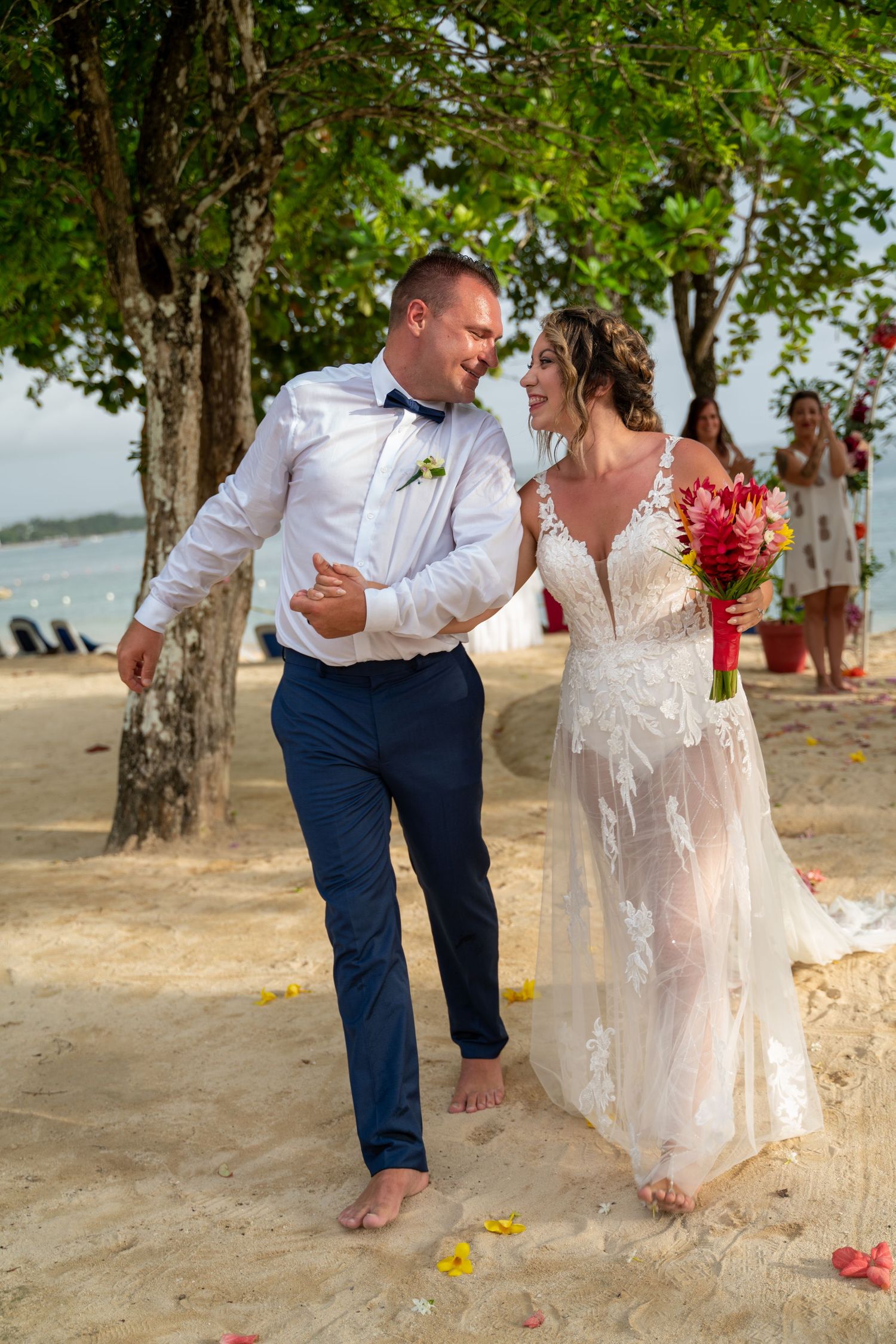 A couple in wedding attire shares playful moments on a sandy beach with trees in the background.