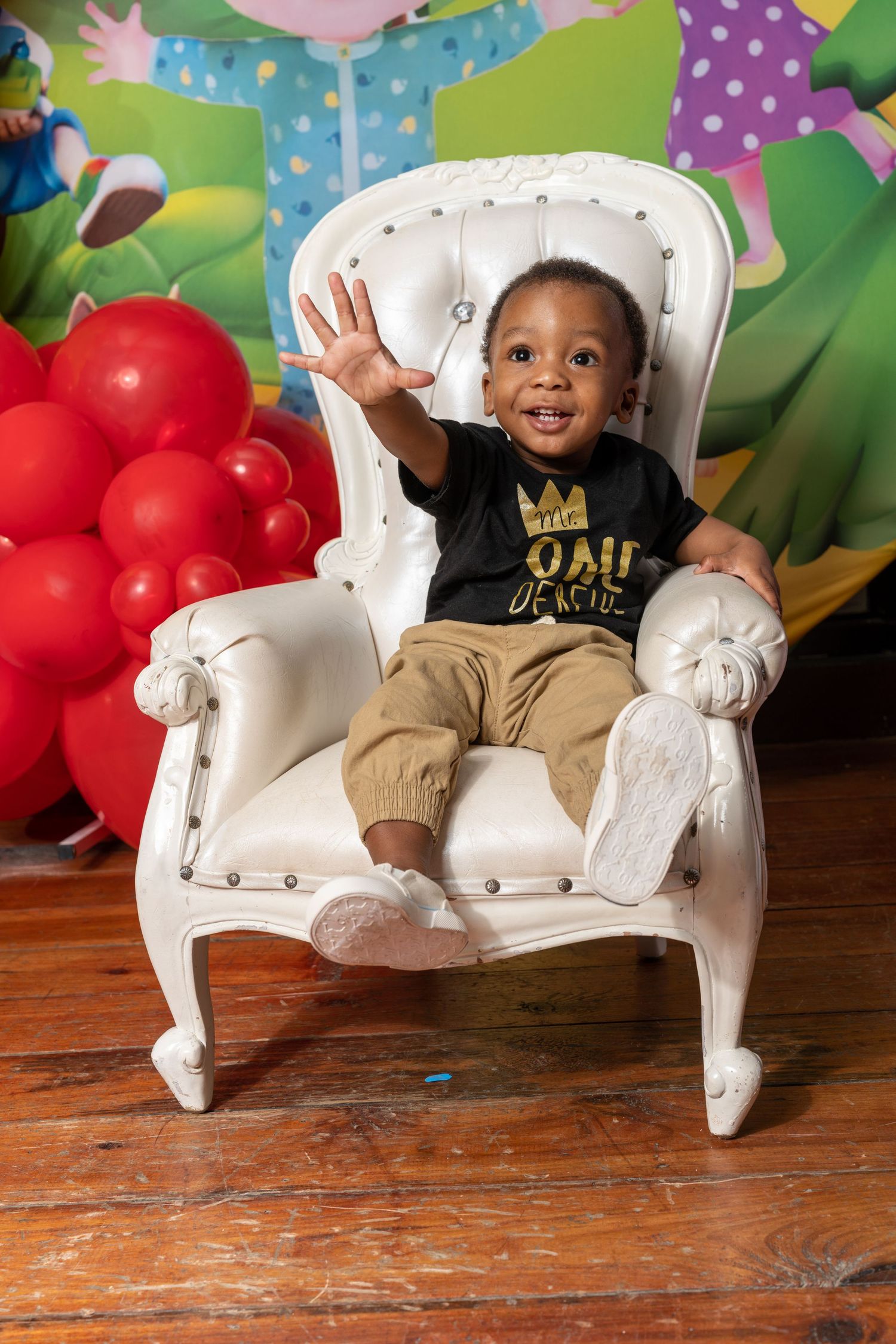 Baby sitting in white ornate chair with red balloons and polka dot backdrop during professional birthday photo session.