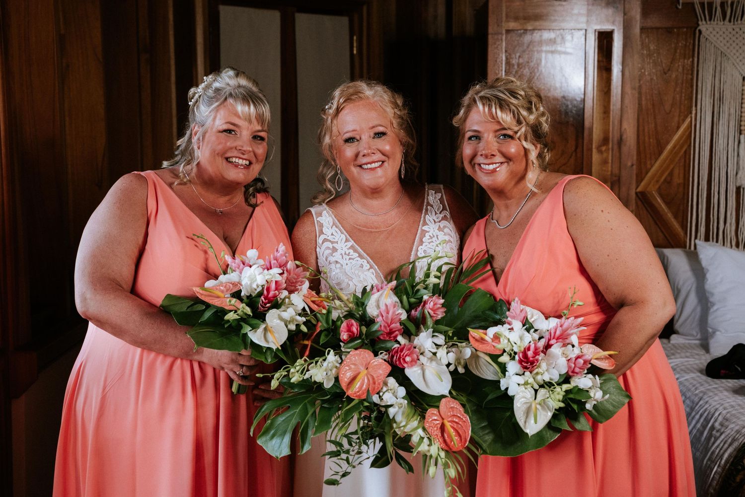 Bridesmaids in coral pink dresses hold beautiful bouquets of white and peach flowers at a rustic venue.