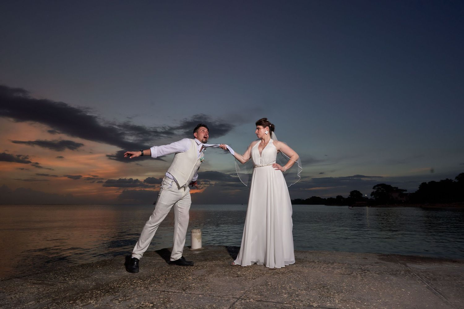 A couple in white attire embraces while standing on a sandy beach at sunset with the ocean in the background.