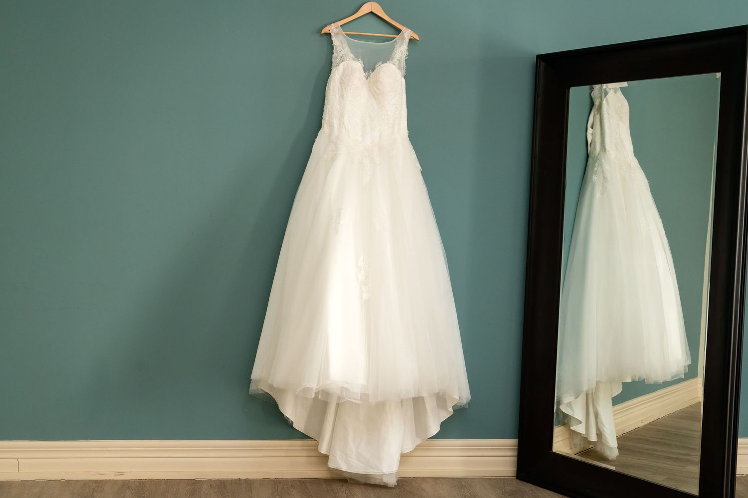 White wedding dress hanging on wooden hanger against mint green wall with decorative mirrors.