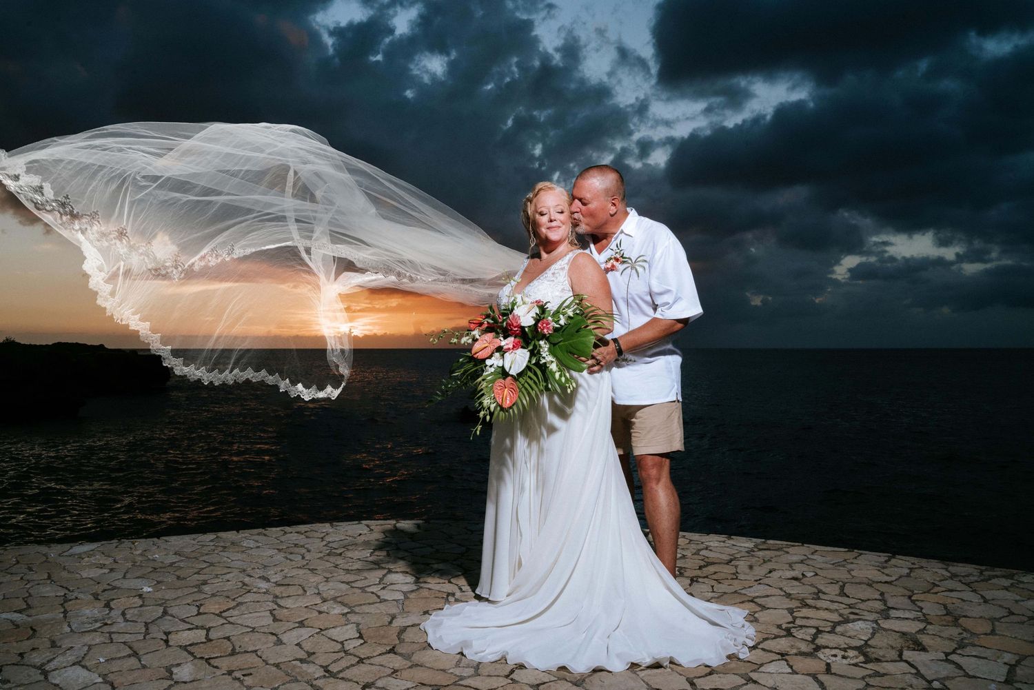 A bride's veil flows dramatically against a dark stormy sky during an outdoor wedding photoshoot.