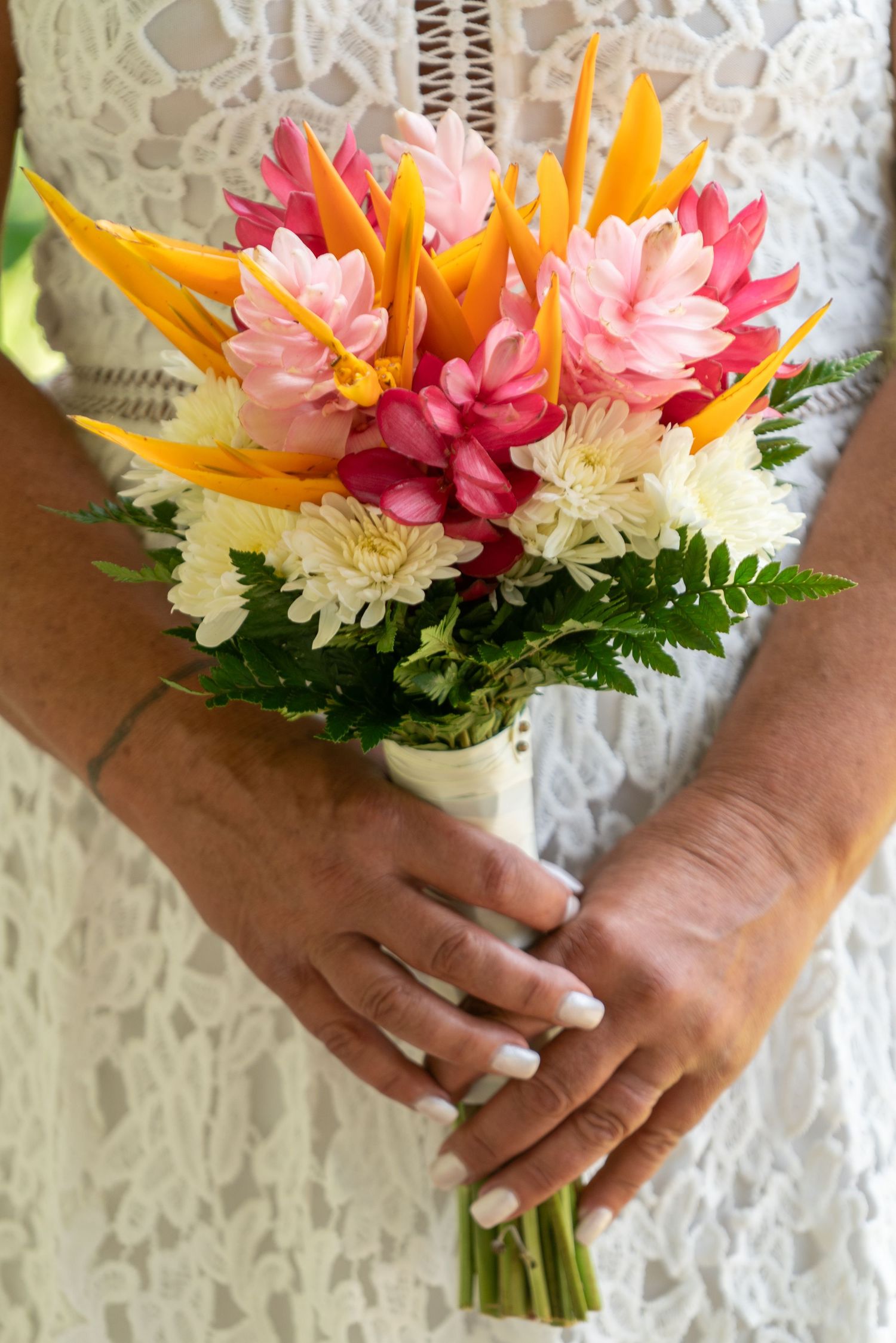Vibrant tropical bridal bouquet with pink ginger flowers, yellow birds of paradise, and white chrysanthemums against lace.