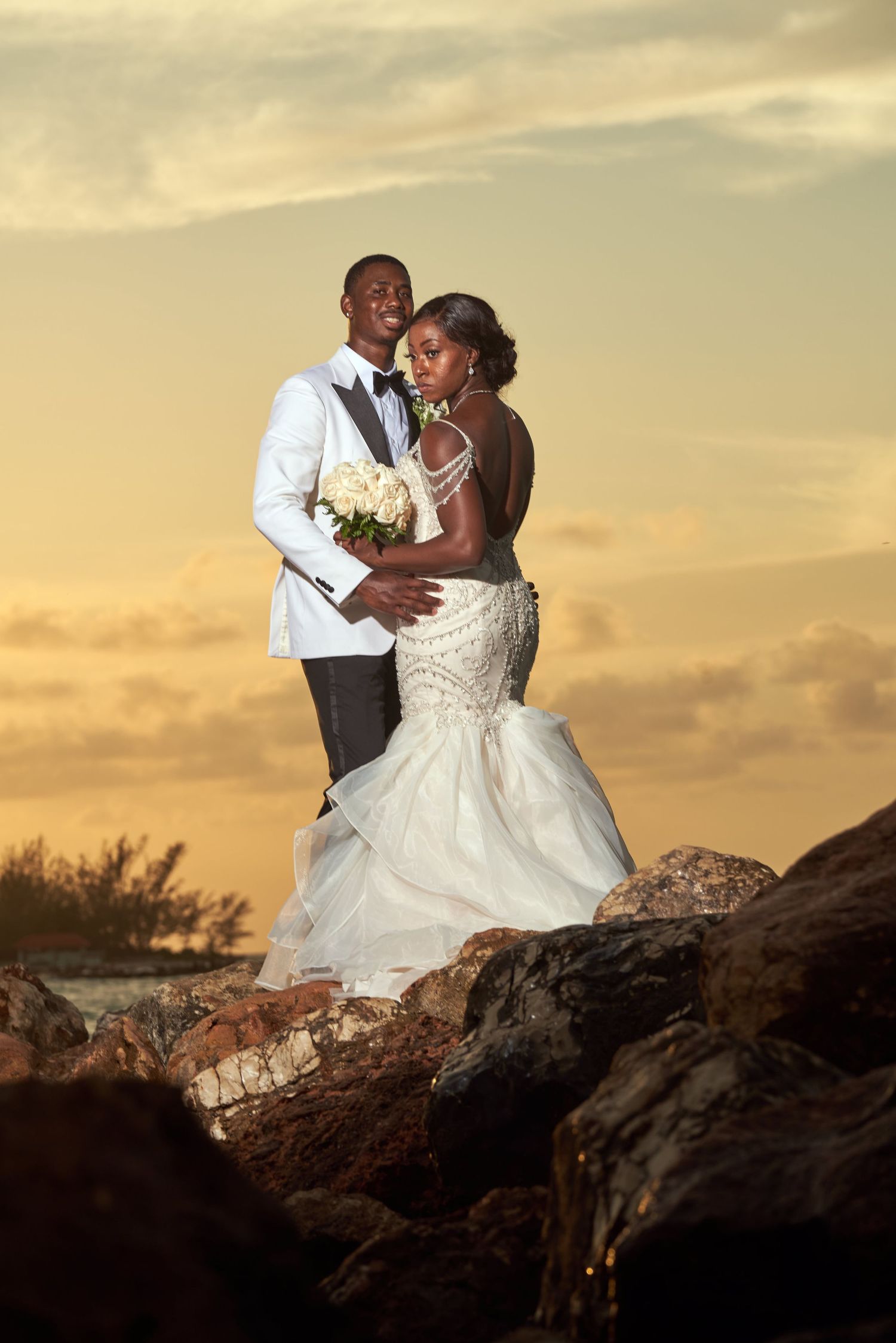 Dramatic sunset wedding photos on rocky beach terrain with couple in elegant attire.