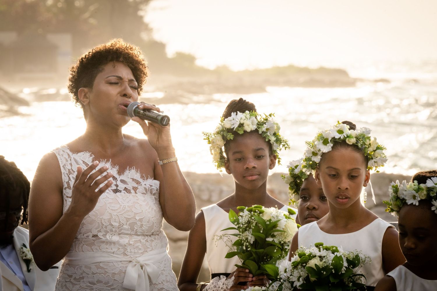 Elegant bridal party in white dresses pose together during golden sunset at tropical beach wedding celebration.